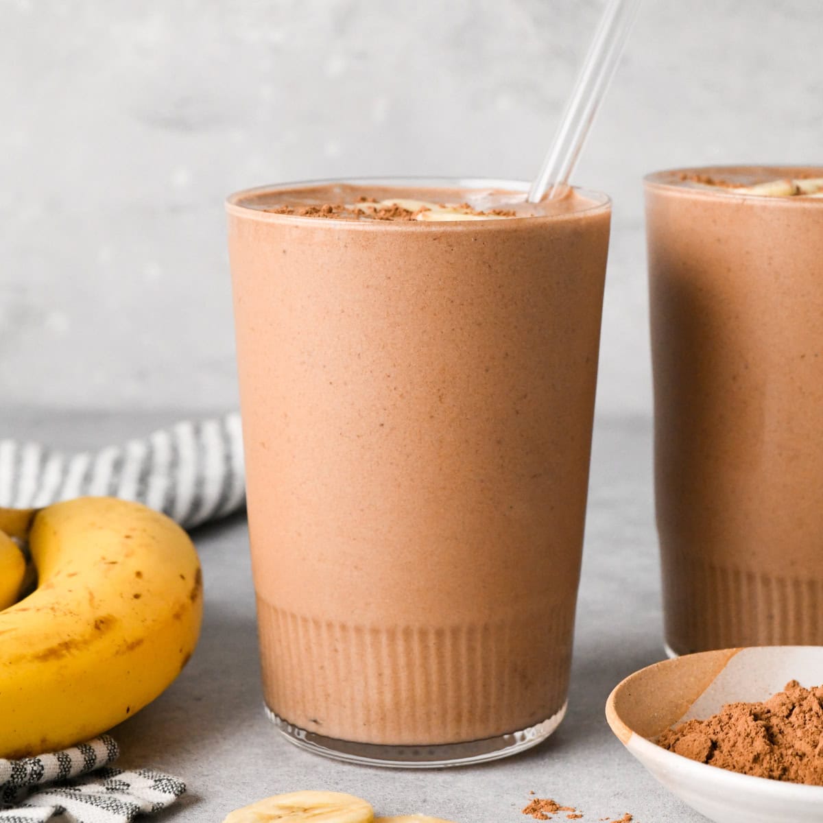 A tall glass of creamy chocolate banana smoothie topped with banana slices and cocoa powder, with a clear straw, fresh bananas, and a small bowl of cocoa powder nearby on a light gray surface.