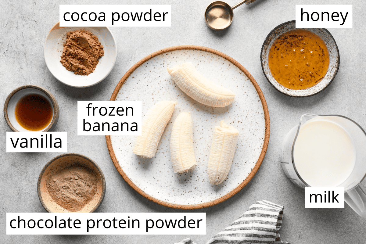 Overhead view of chocolate banana smoothie ingredients arranged on a gray surface, including frozen bananas on a plate, cocoa powder, chocolate protein powder, vanilla extract, honey, and milk in small bowls and a measuring jug, with each ingredient labeled.