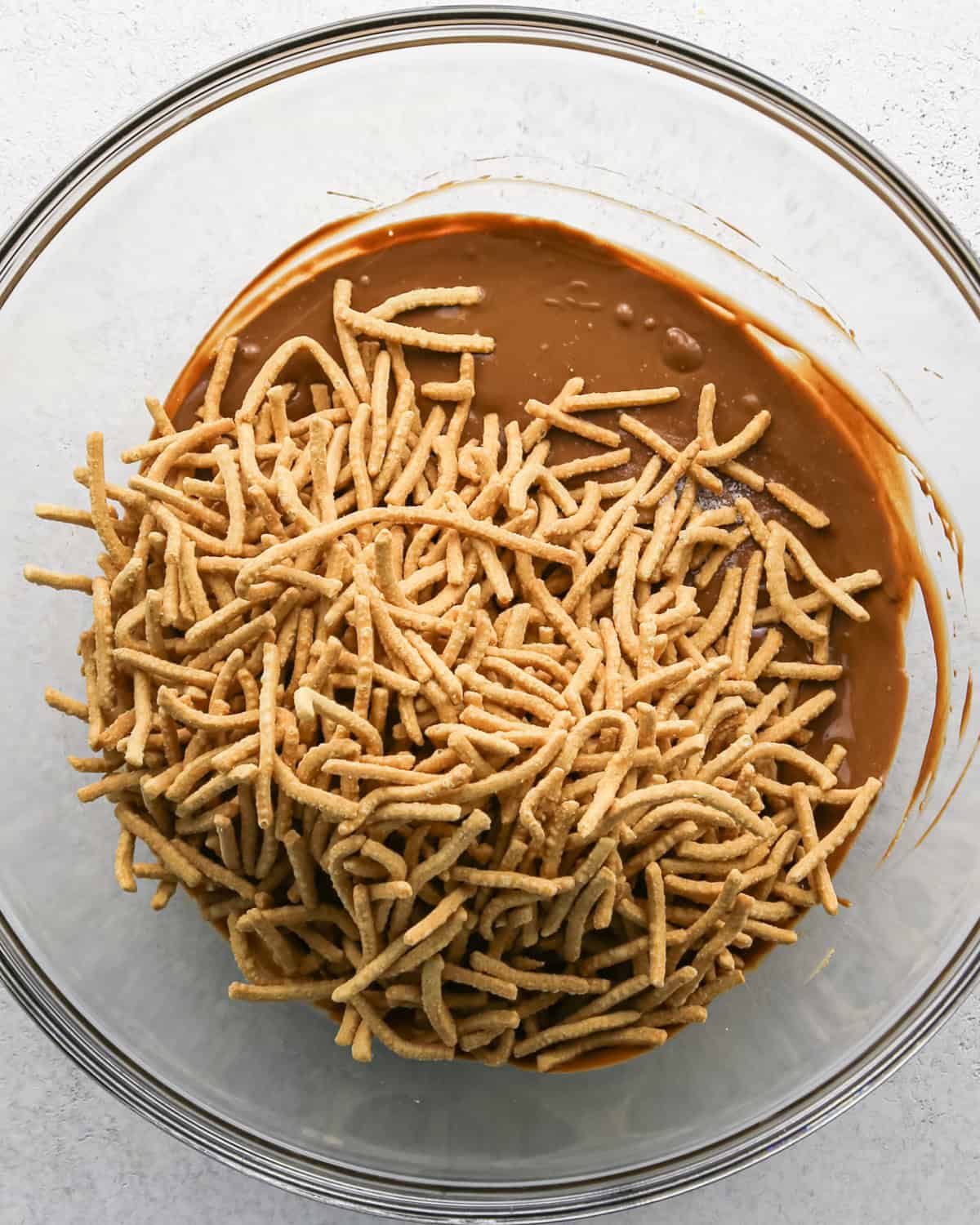 Making Haystack Cookies: overhead photo of chow mein noodles added to melted mixture of peanut butter, semisweet chocolate chips, and butterscotch chips in a large clear bowl.