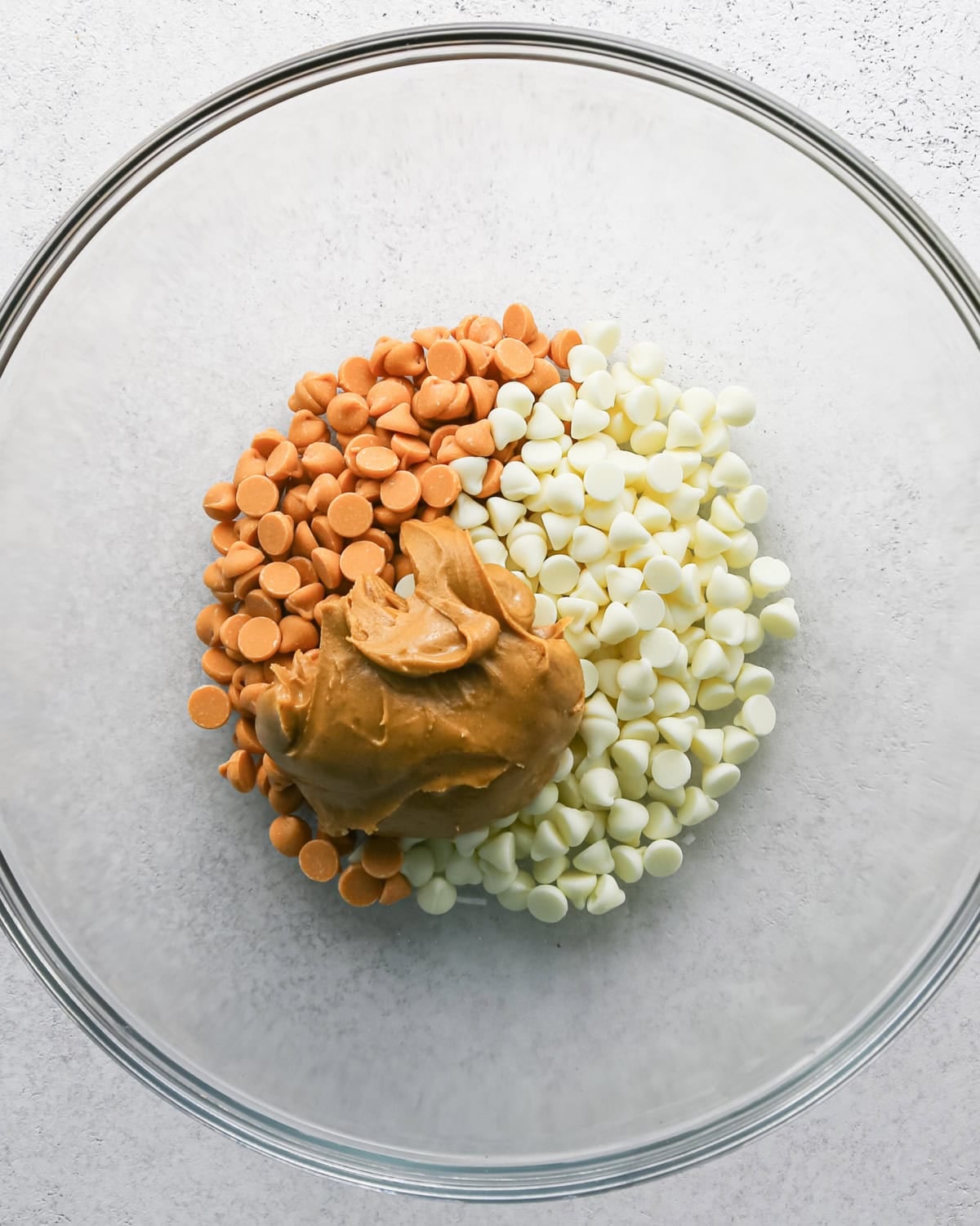Making Haystack Cookies: overhead photo of white chocolate chips, butterscotch chips, and peanut butter added to a large clear bowl.