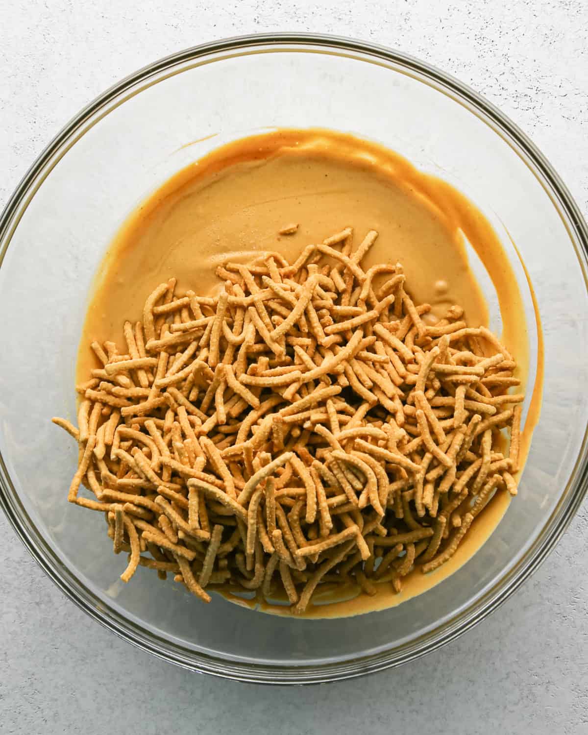 Making Haystack Cookies: overhead photo of chow mein noodles added to melted mixture of peanut butter, white chocolate chips, and butterscotch chips in a large clear bowl.