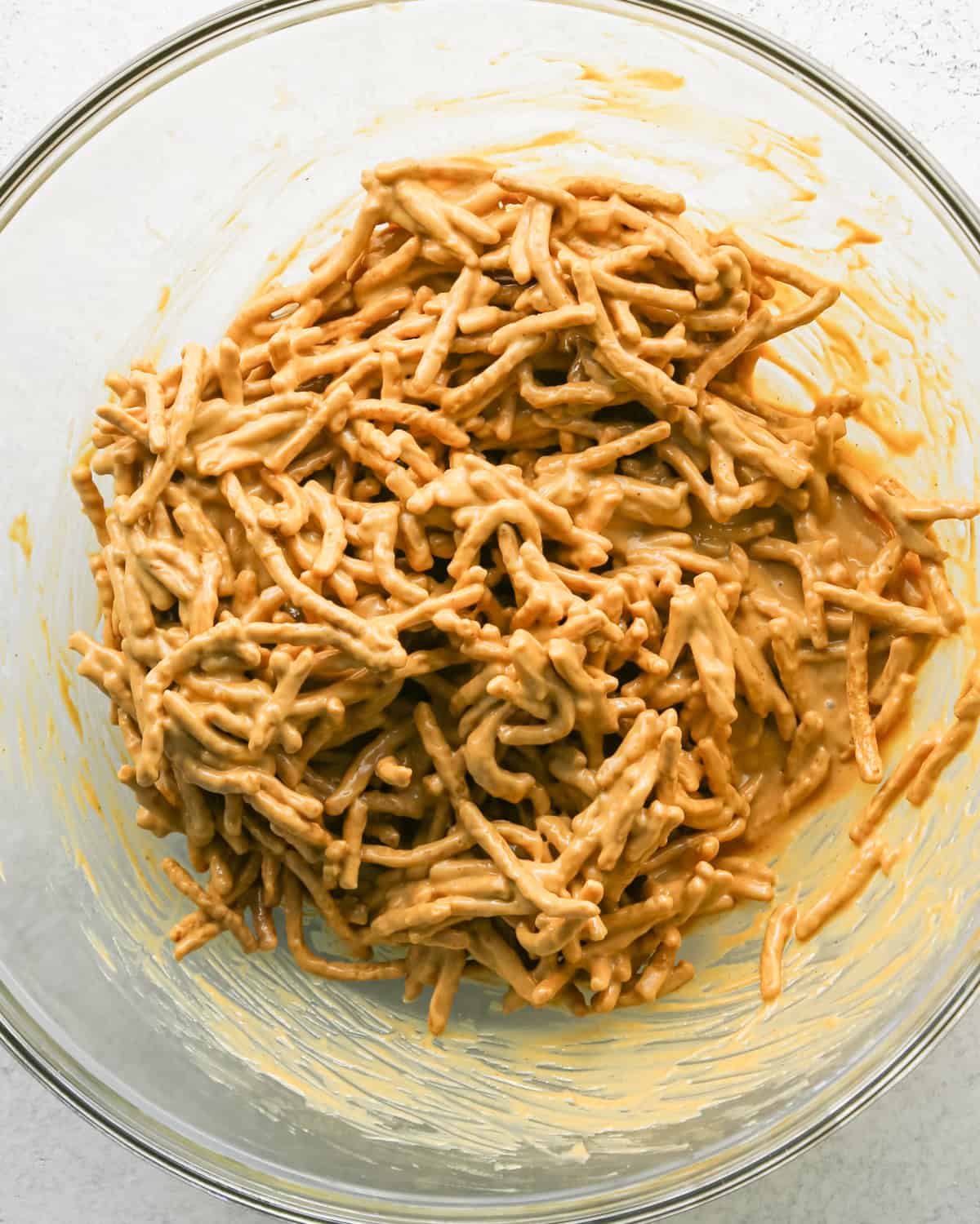 Making Haystack Cookies: overhead photo of chow mein noodles added to melted mixture of peanut butter, white chocolate chips, and butterscotch chips in a large clear bowl and mixed together.