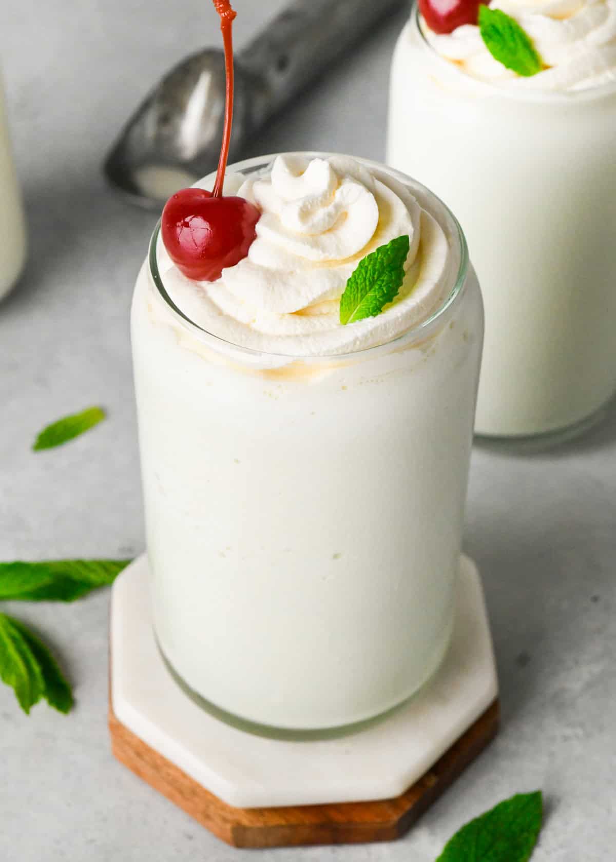 two glasses filled with Shamrock Shake both topped with whipped cream, fresh mint leaves, and a maraschino cherry. with fresh. mint leaves and an ice cream scoop in the background