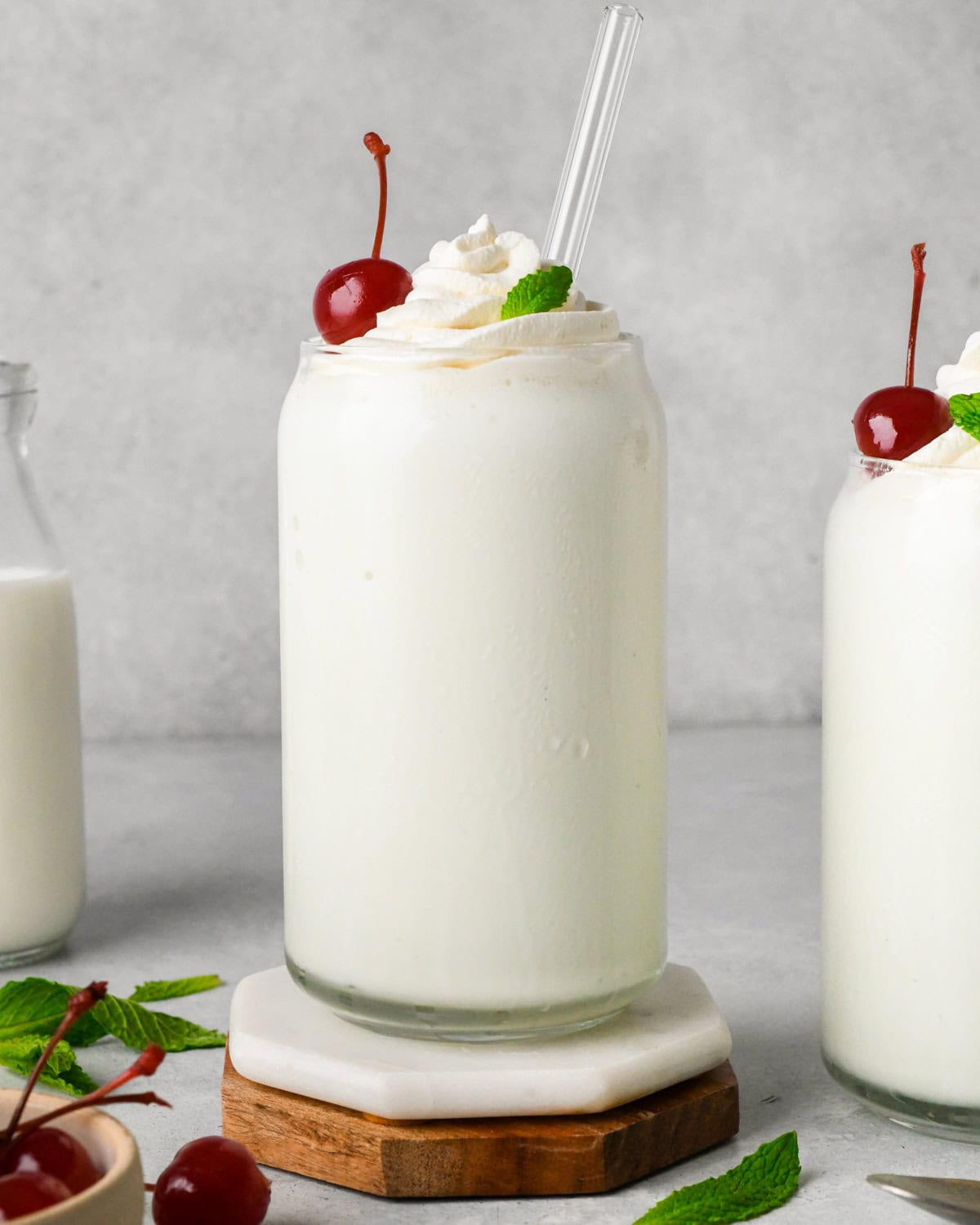 two tall glasses filled with Shamrock Shake both topped with whipped cream, fresh mint leaves, and a maraschino cherry. Surrounded by cherries, mint leaves and a glass of milk.