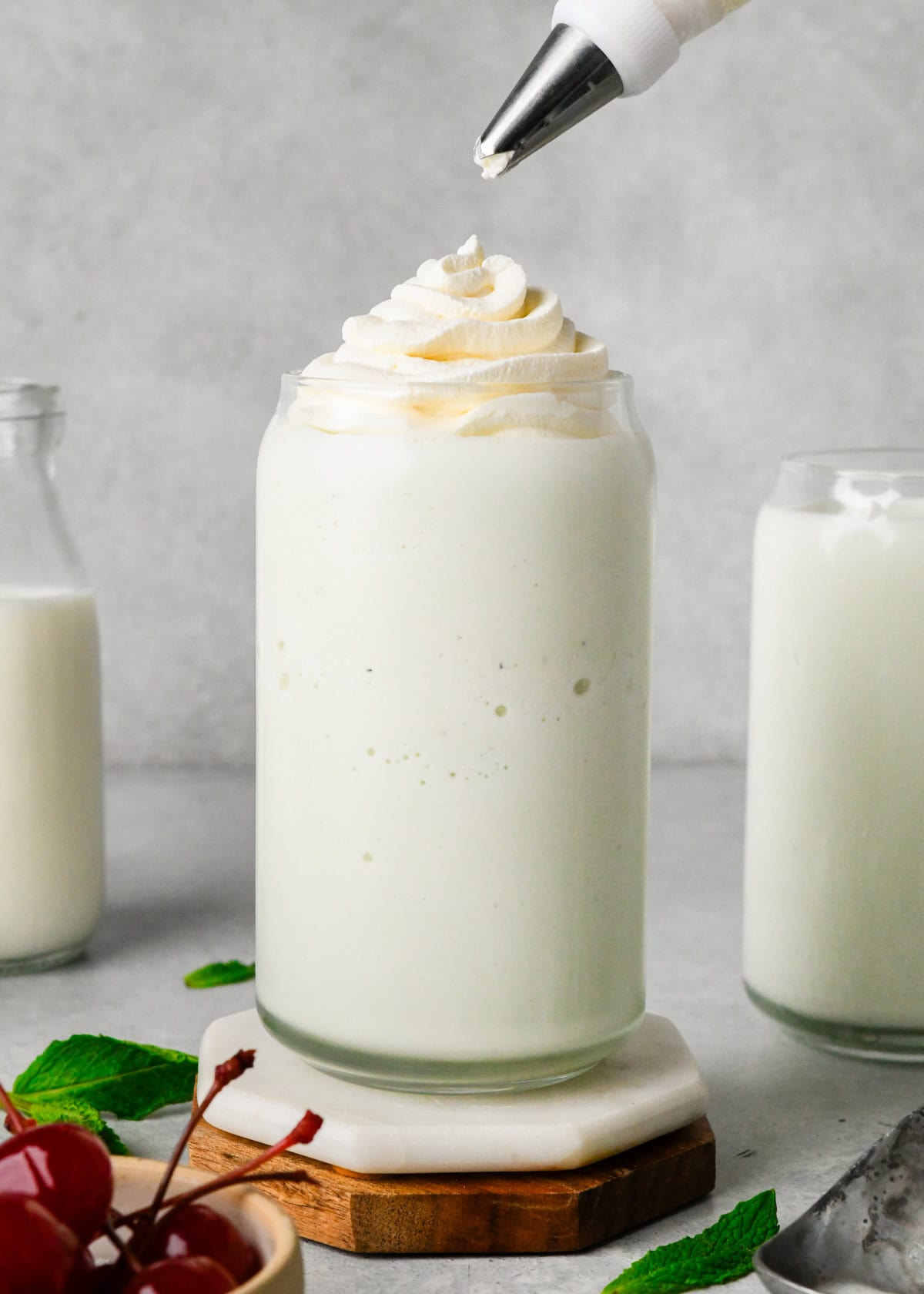 whipped cream being piped onto a shamrock shake in a glass surrounded by another glass of shamrock shake, cherries and mint leaves.