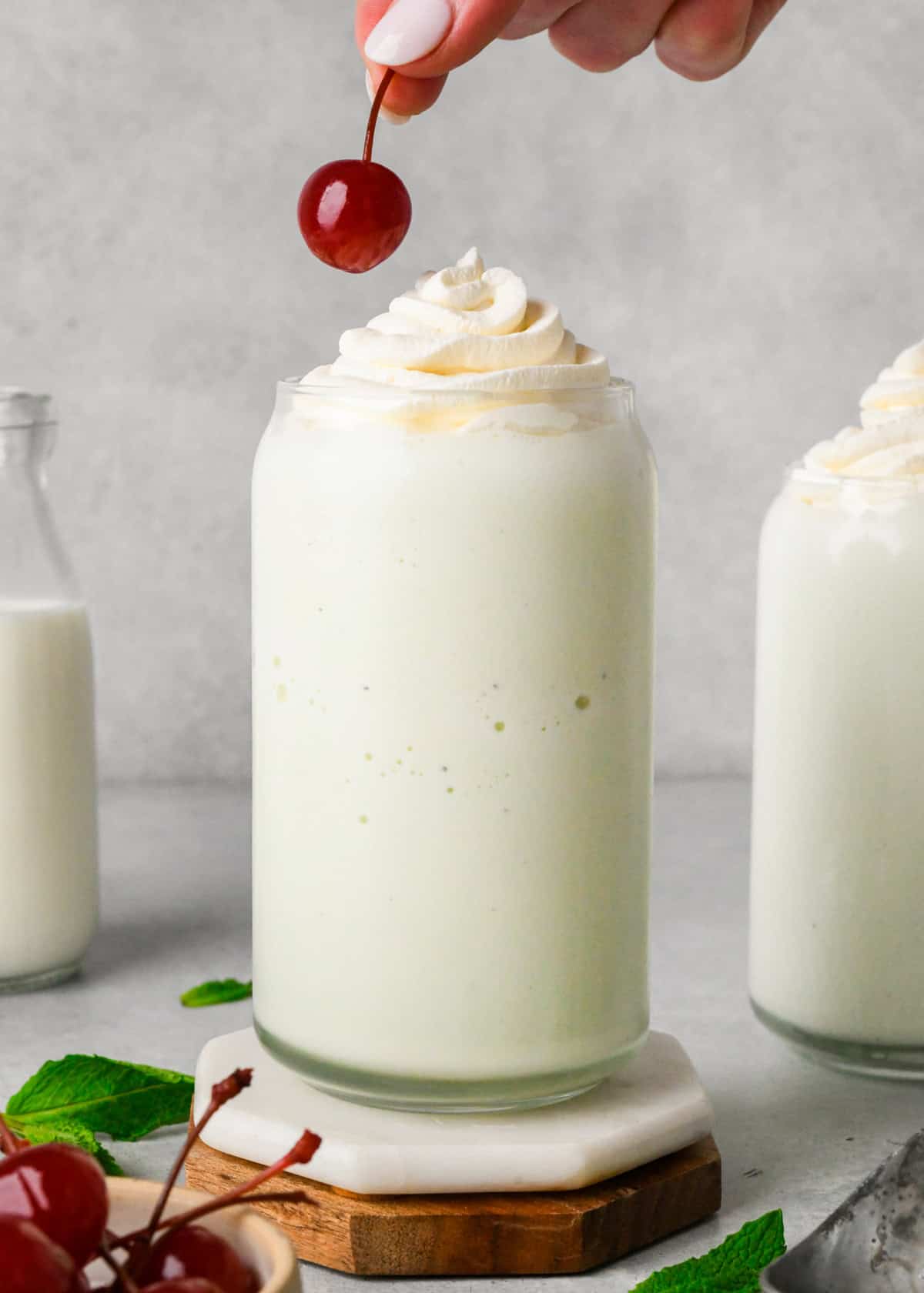 a cherry being put onto a shamrock shake in a glass topped with whipped cream surrounded by another glass of shamrock shake, cherries and mint leaves.