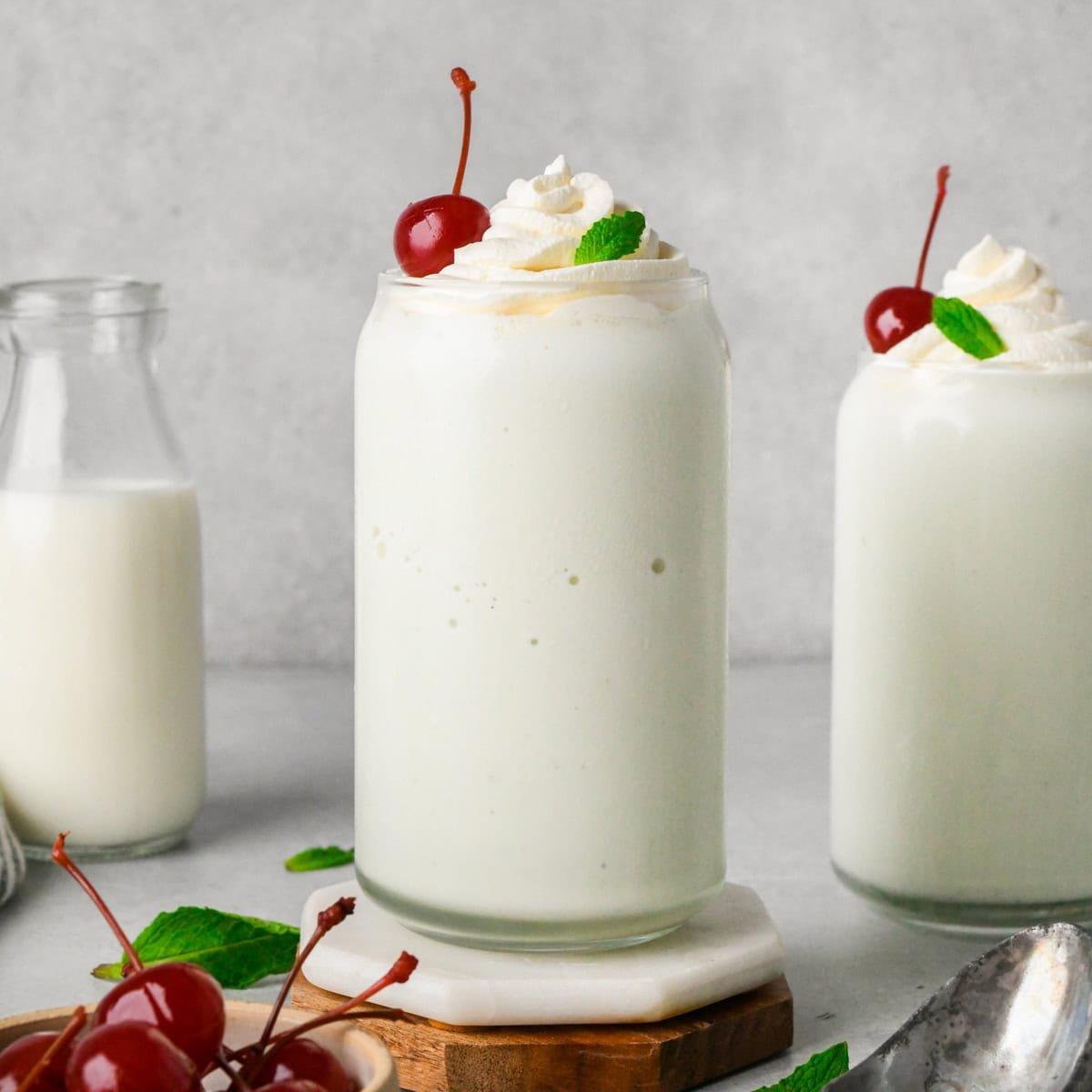 two tall glasses filled with Shamrock Shake both topped with whipped cream, fresh mint leaves, and a maraschino cherry. Surrounded by cherries, mint leaves and a glass of milk.