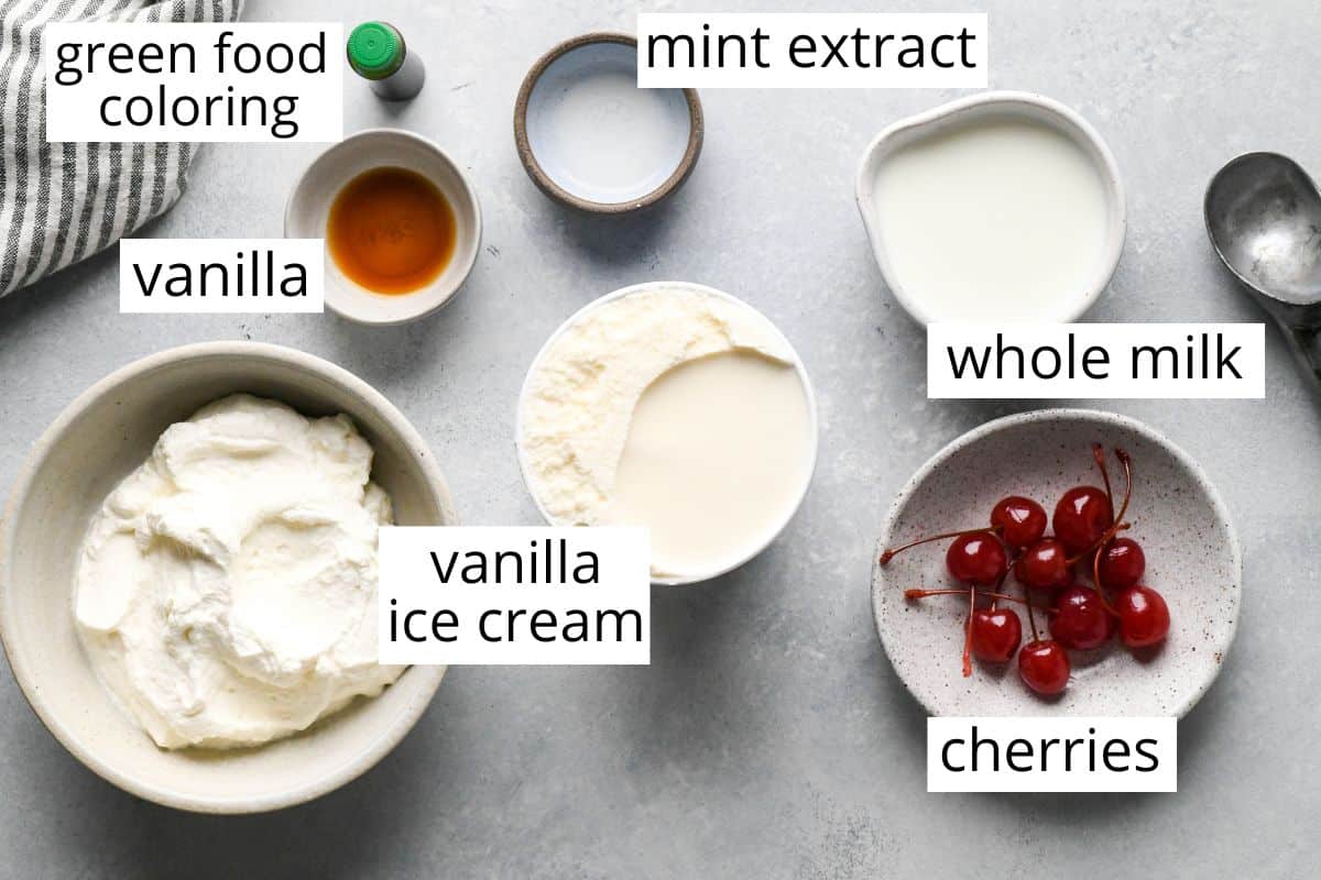 overhead photo of the labeled ingredients in this shamrock shake recipe like vanilla, food coloring, mint extract, vanilla ice cream, whole milk and cherries.