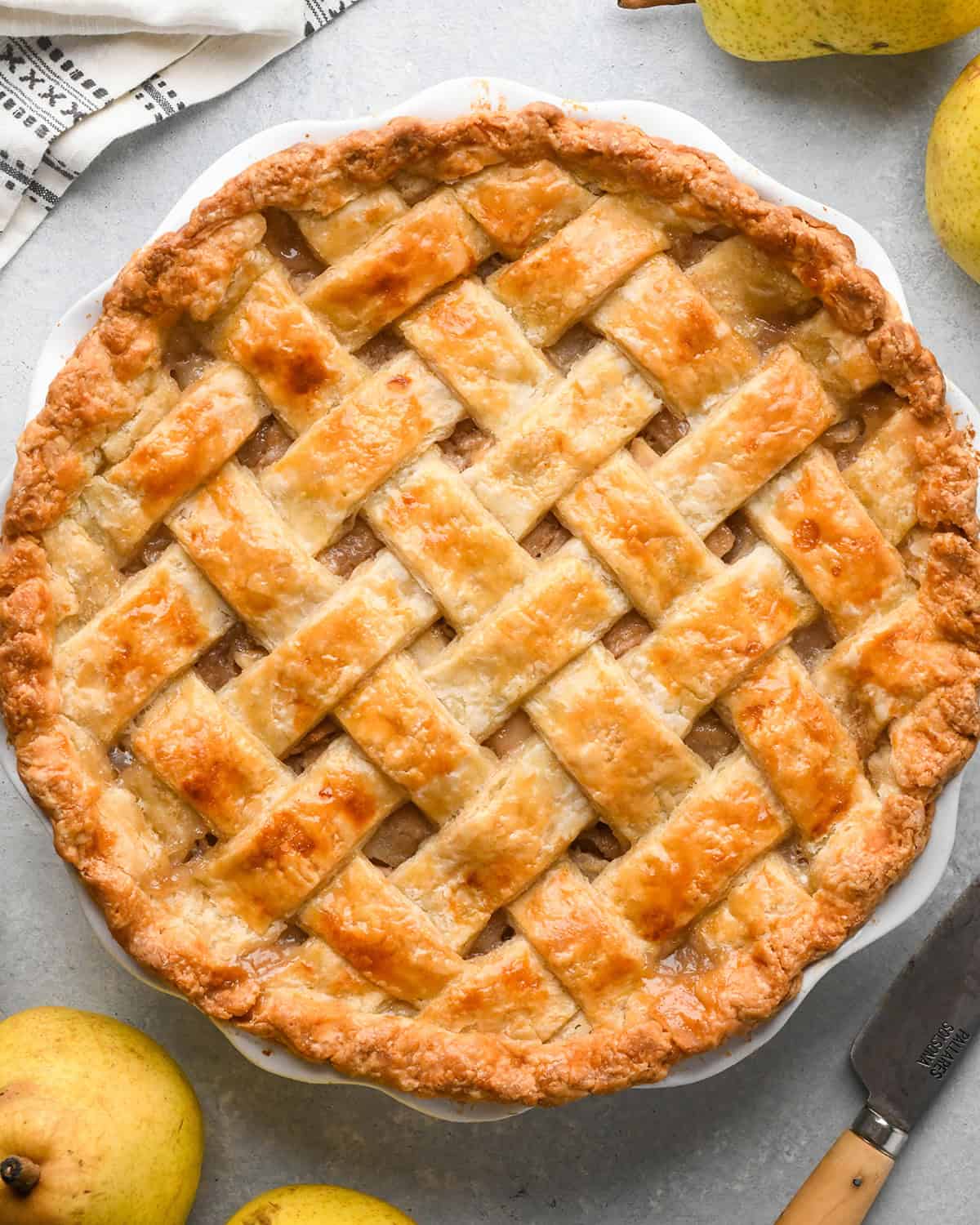 pear pie with a lattice crust in a white pie dish on a gray surface with pears in the bottom left and top right corners
