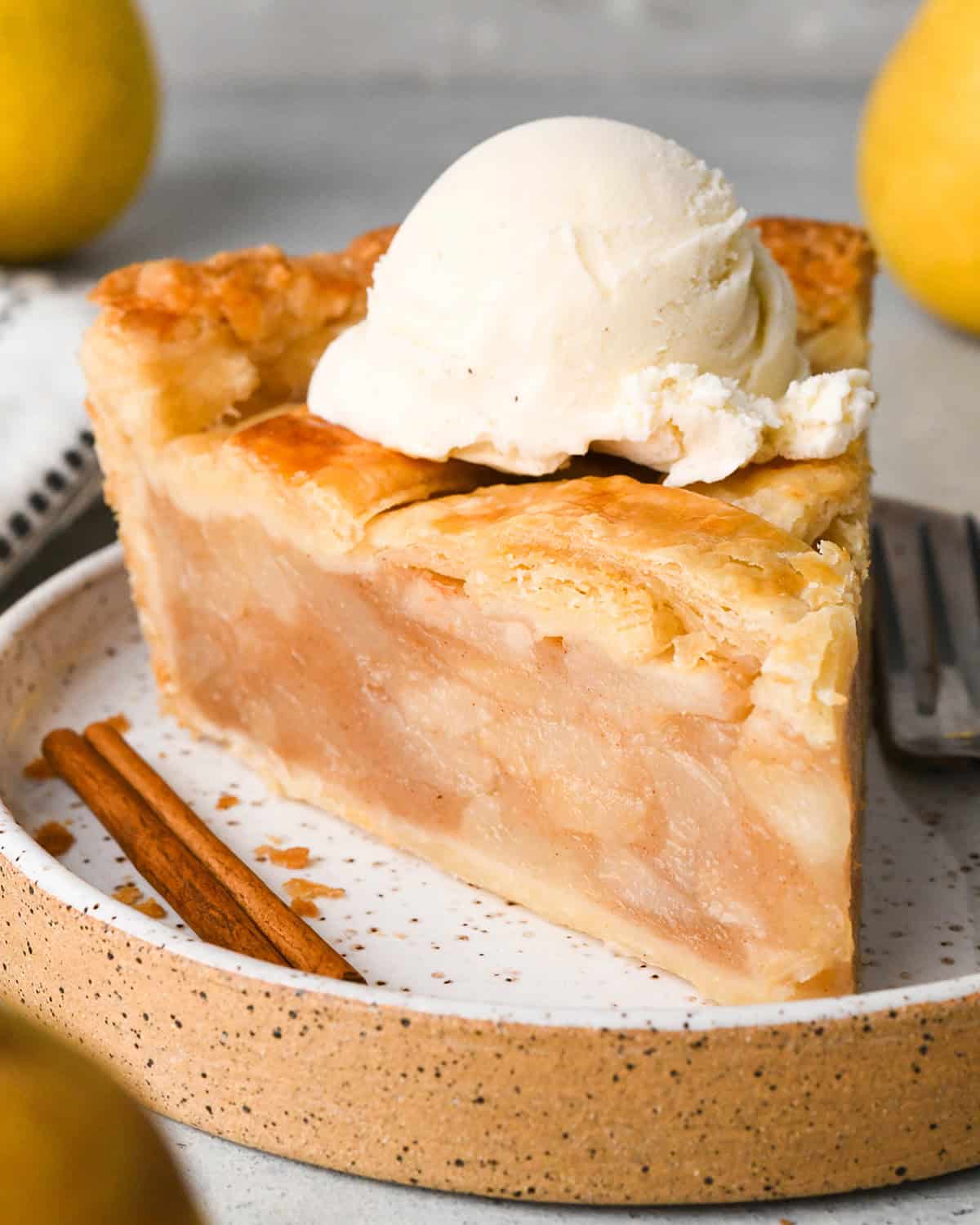 a slice of pear pie with vanilla ice cream on top on a plate with a cinnamon stick and a fork and pears in the background. 