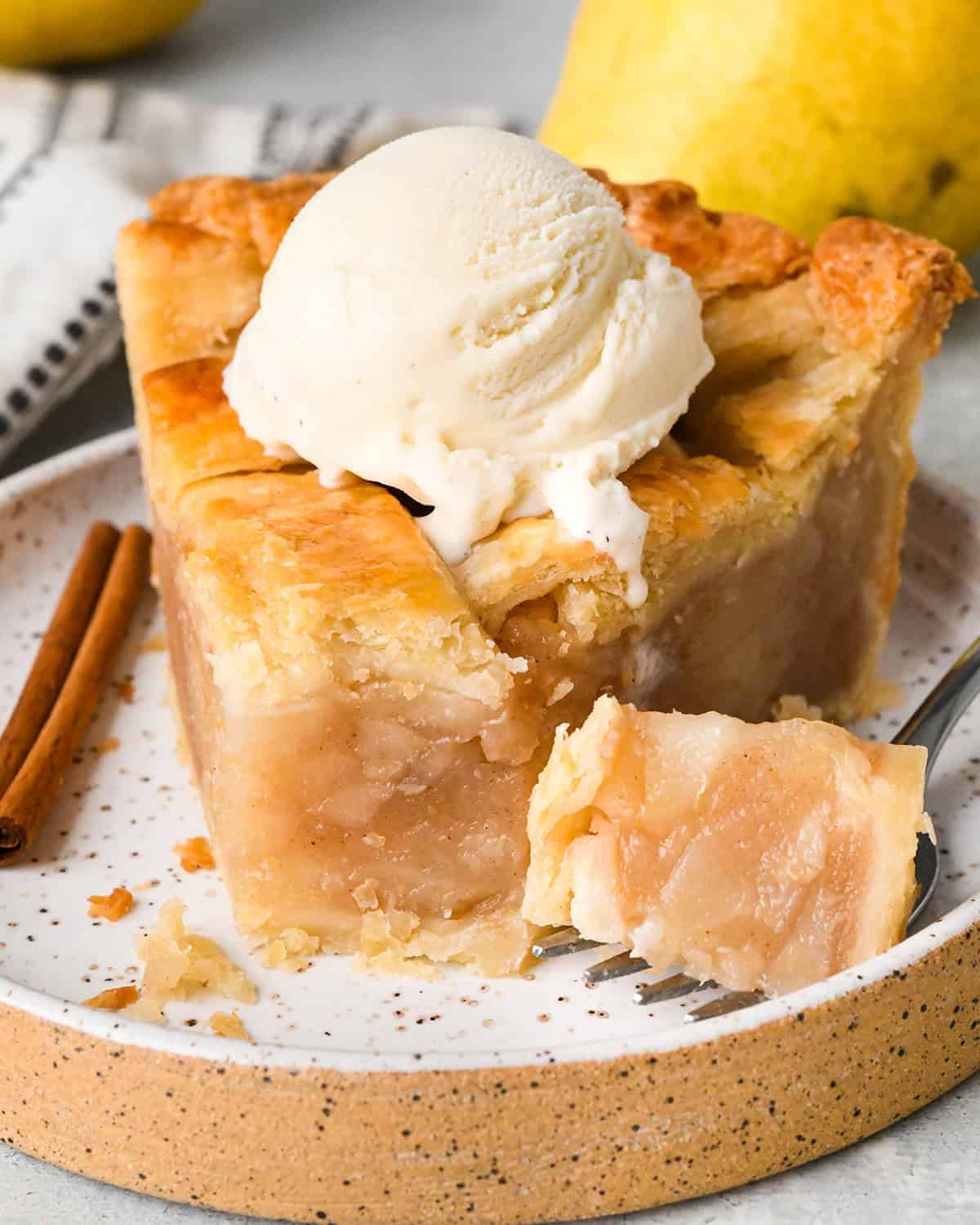 a slice of pear pie with vanilla ice cream on top on a plate with a cinnamon stick and a fork with a bite on it and pears in the background. 