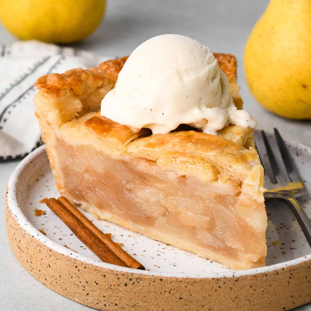 a slice of pear pie with vanilla ice cream on top on a plate with a cinnamon stick and a fork and pears in the background. 