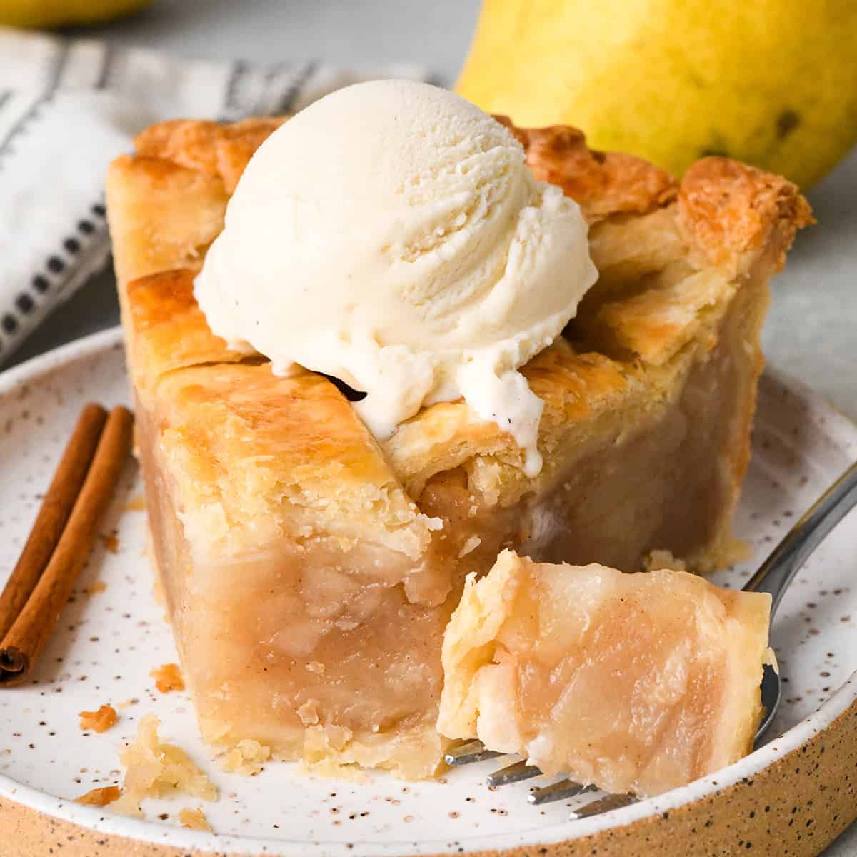 a slice of pear pie with vanilla ice cream on top on a plate with a cinnamon stick and a fork with a bite on it and a pear in the background. 