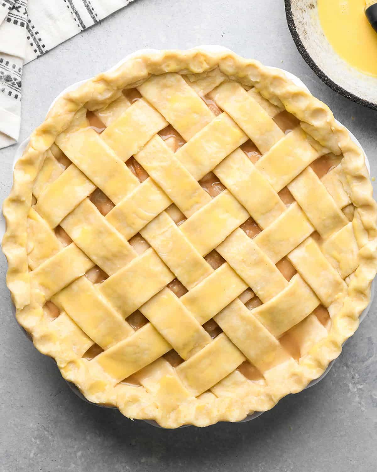 lattice pie crust brushed with egg wash on a gray surface with a black and white towel in the upper left corner