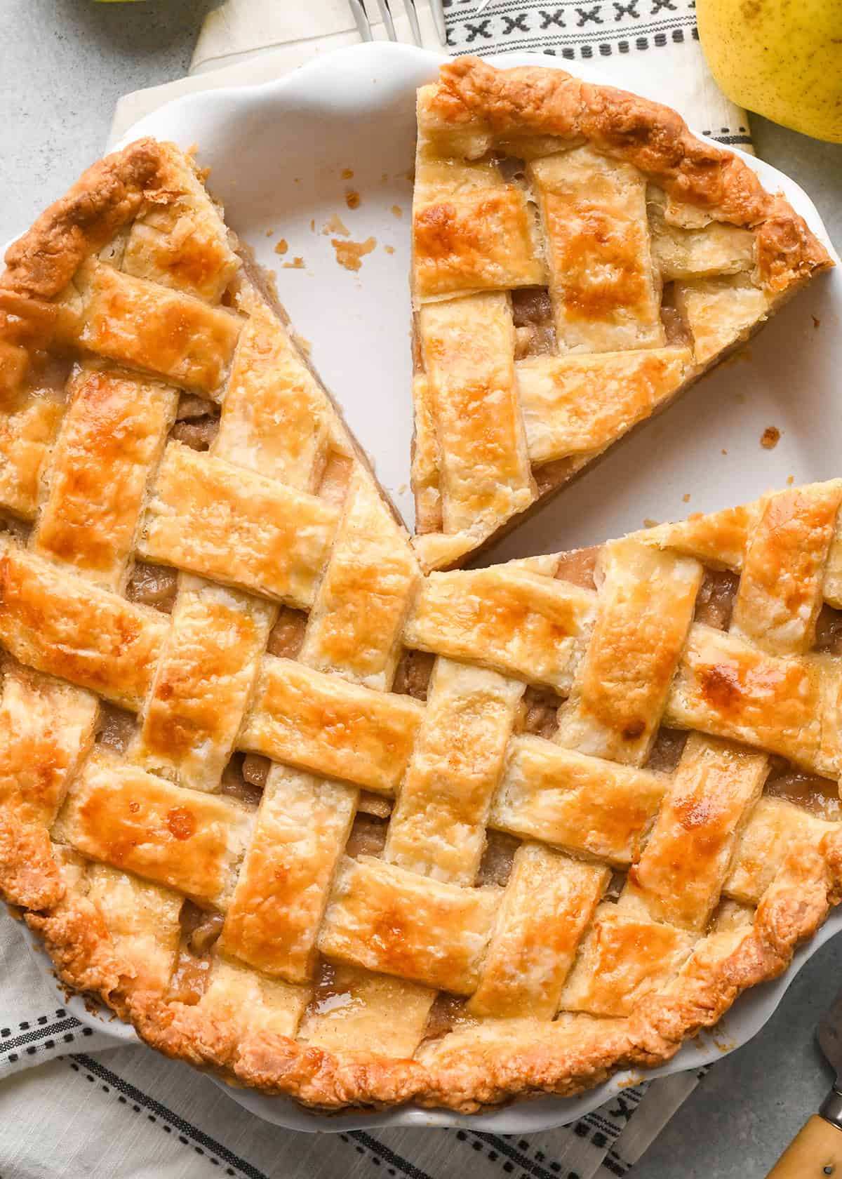 a pie with a Lattice Pie Crust and a piece cut out of it in a pie dish on a gray surface with a black and white towel