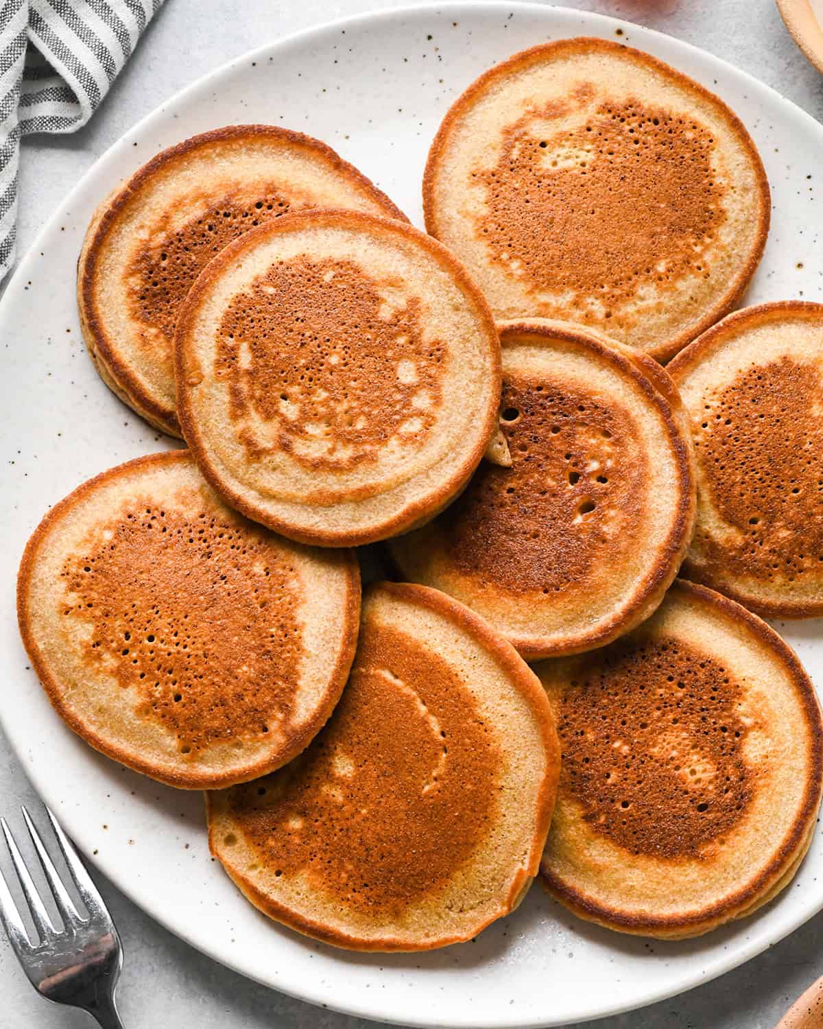 8 oat flour pancakes on a plate on a gray surface with a striped towel in the top left corner and a fork in the bottom left corner