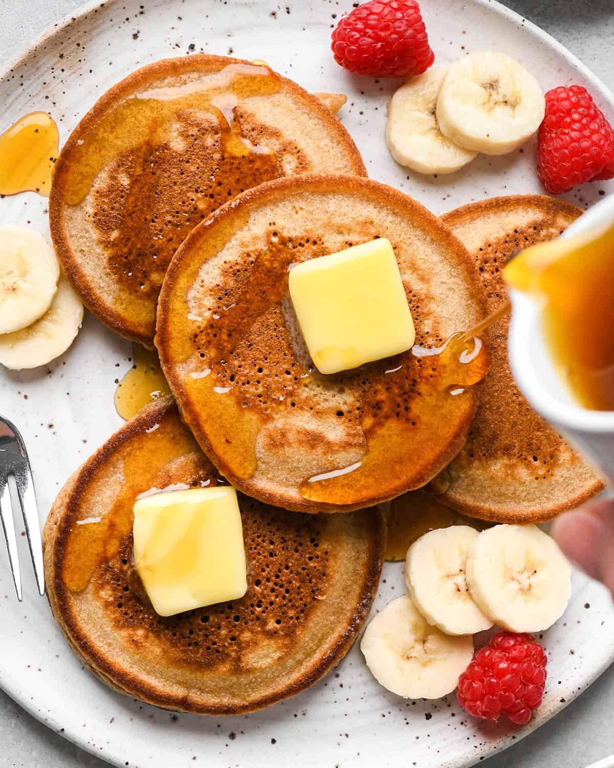 syrup being poured over 4 oat flour pancakes on a white plate with butter on top of two pancakes and bananas and raspberries on the plate surrounding the pancakes