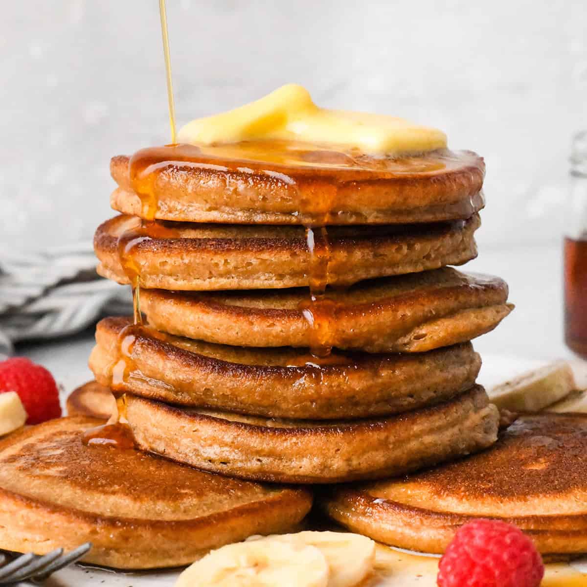 a stack of 7 oat flour pancakes topped with melted butter and maple syrup being poured over the stack. Bananas and raspberries on the plate.