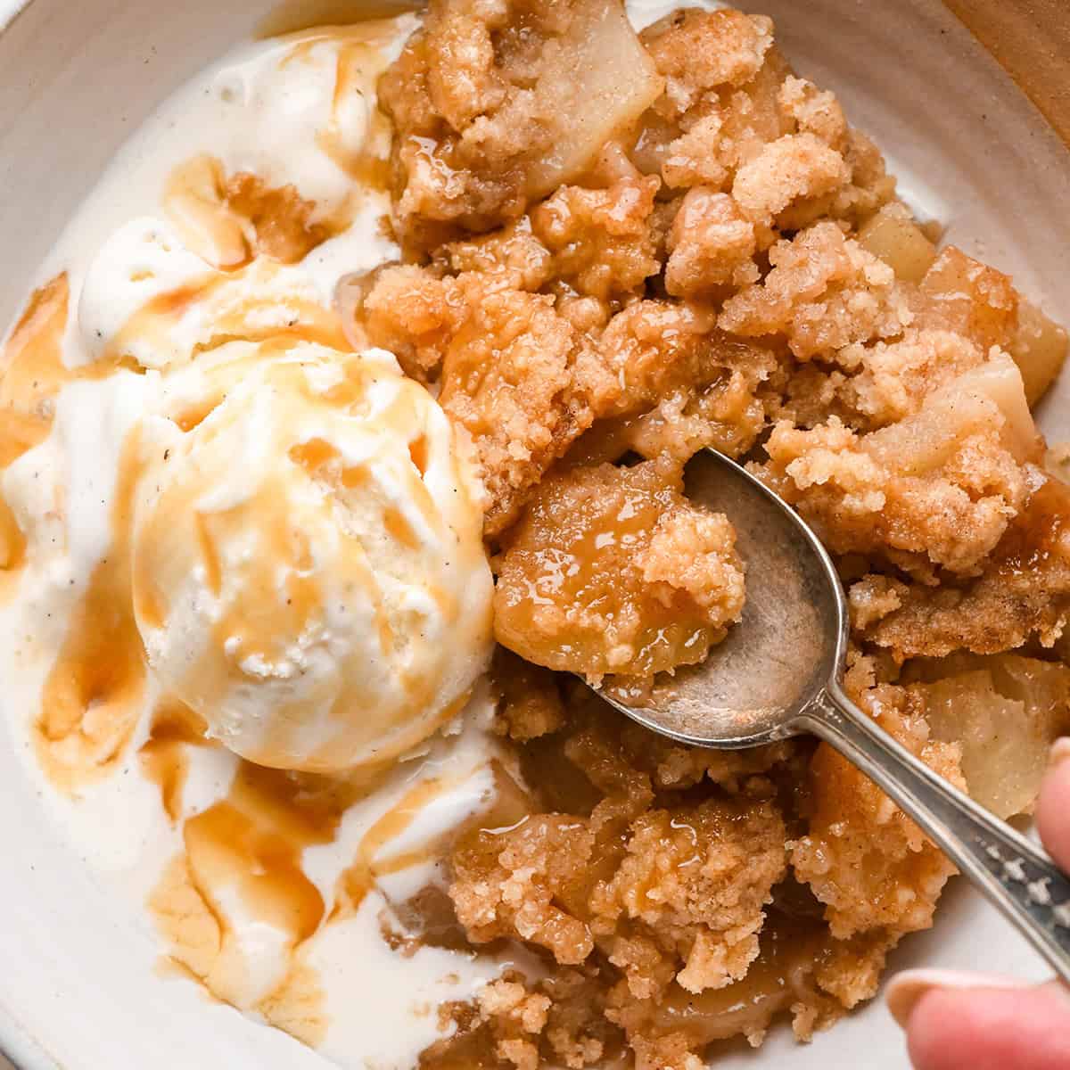 a serving of Pear Crumble in a bowl with a spoon, vanilla ice cream and caramel sauce