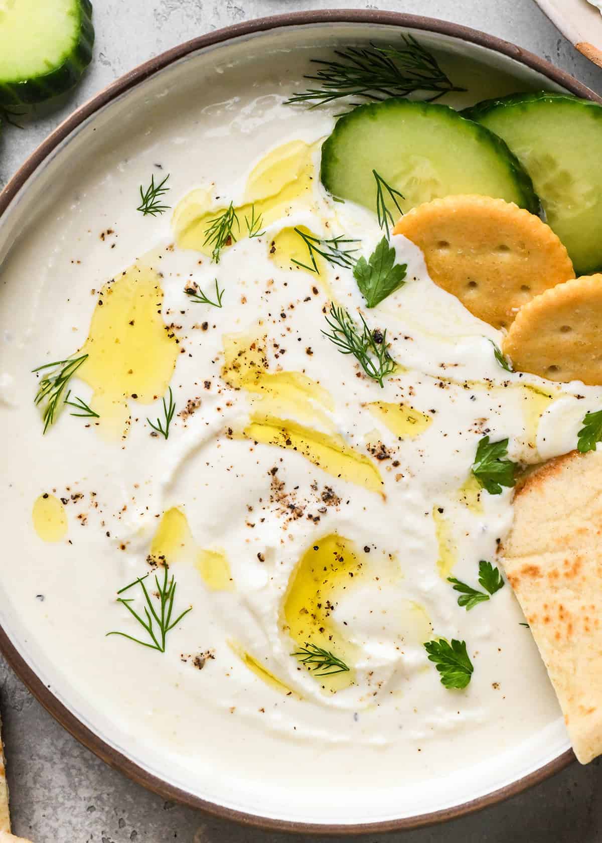 close up of a piece of pita bread being dipped into whipped Goat Cheese Dip in a bowl with crackers herbs oil and cucumbers.
