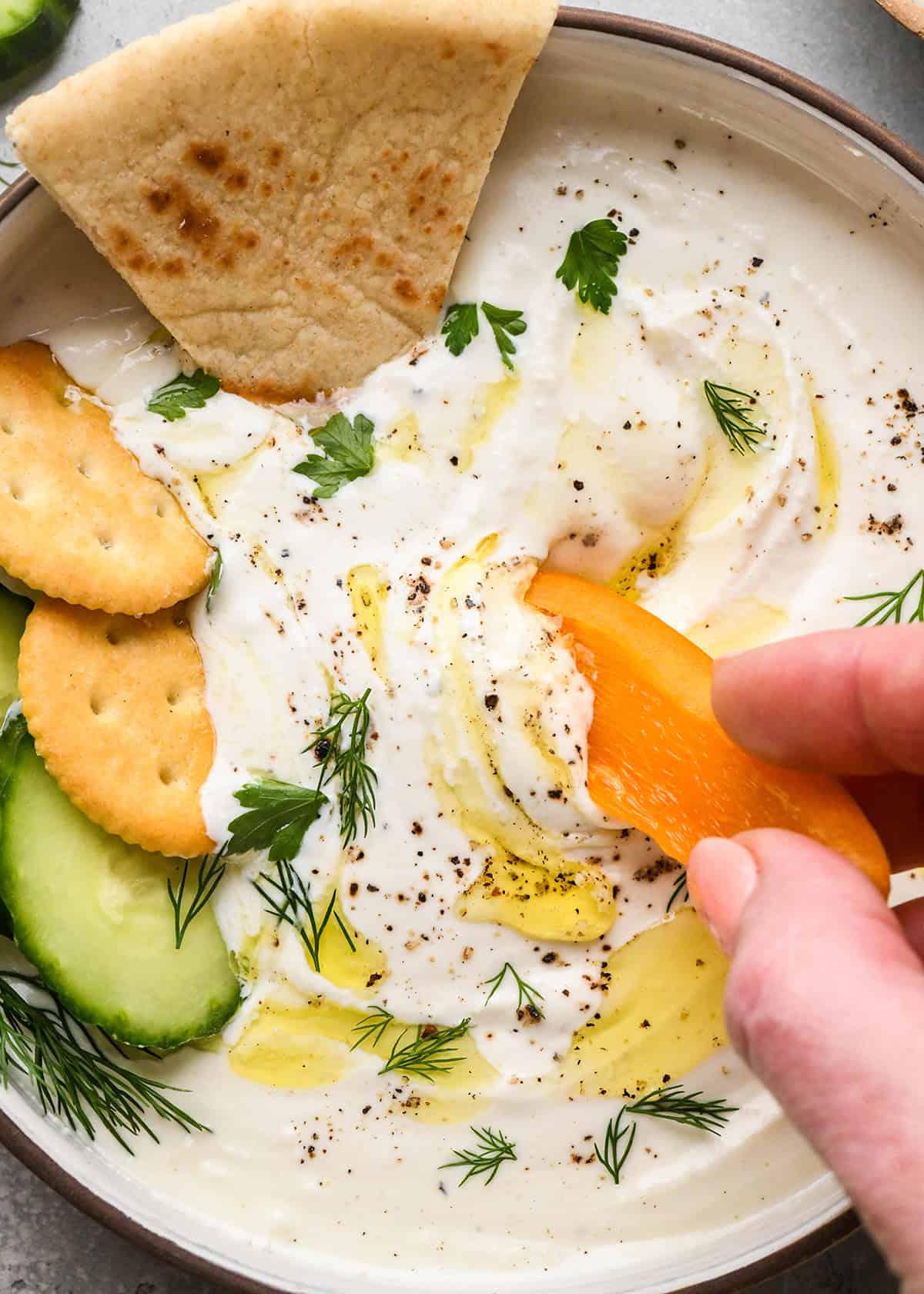 an orange bell pepper being dipped into Whipped Goat Cheese Dip in a bowl garnished with oil, pepper and herbs with crackers and cucumbers in the bowl too