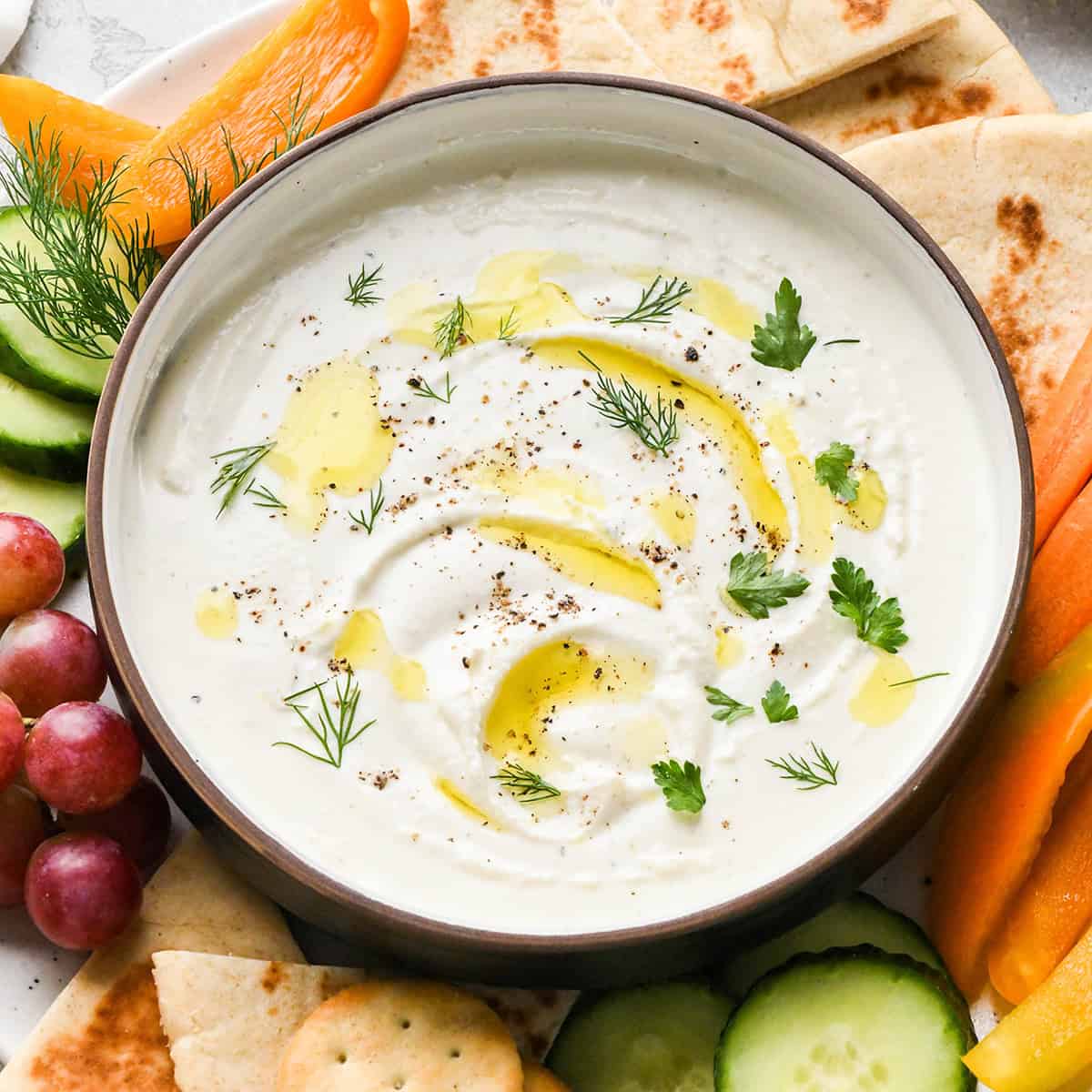 Whipped Goat Cheese Dip in a bowl garnished with oil and herbs surrounded by vegetables, fruit, pita bread and crackers. 