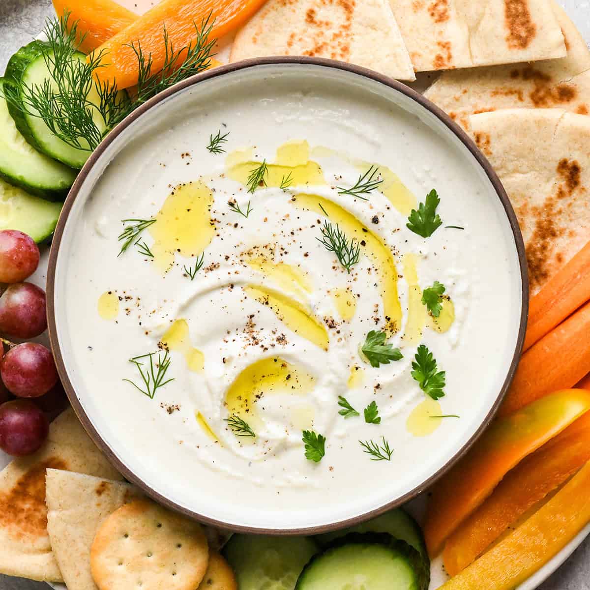 Whipped Goat Cheese Dip in a bowl garnished with oil, salt and pepper and herbs surrounded by cucumbers, bell peppers, pita bread, grapes and crackers
