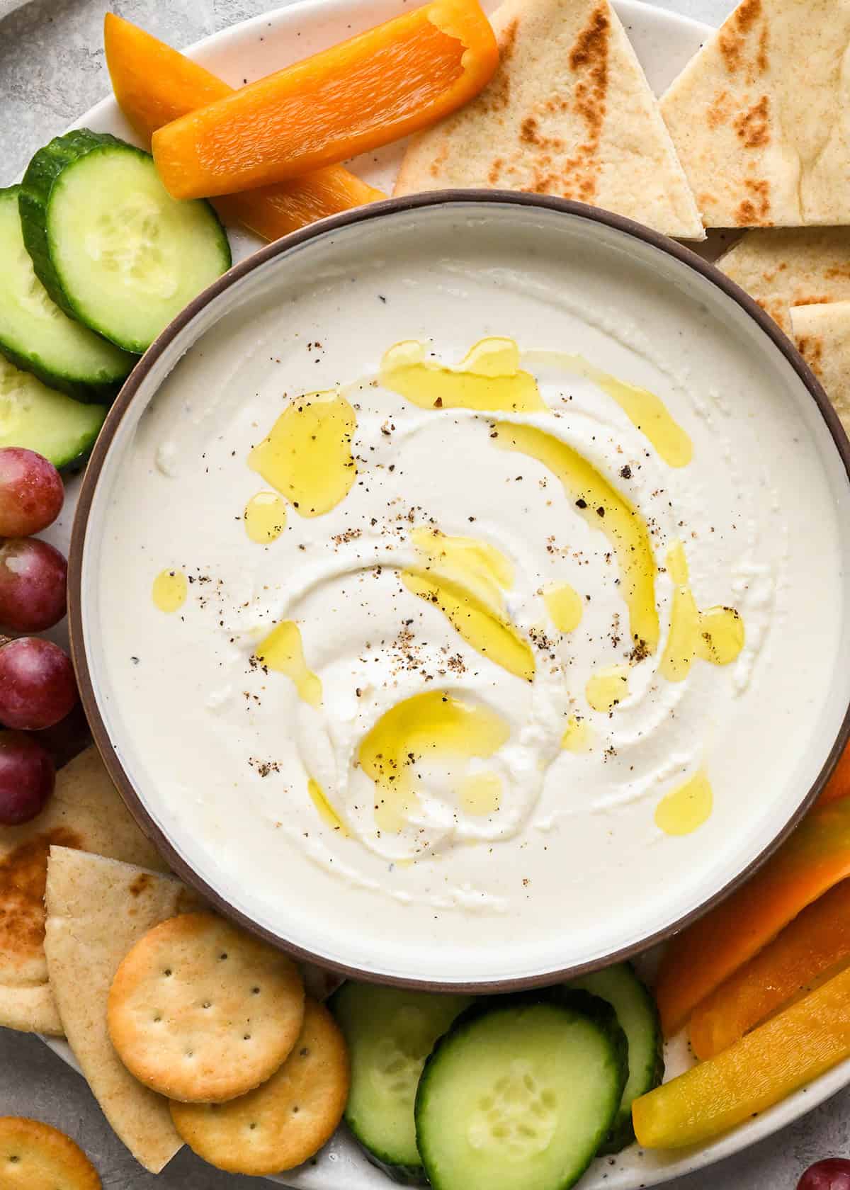 Whipped Goat Cheese Dip in a serving bowl with oil and pepper on top, surrounded by pita bread, cucumbers, grapes, crackers and peppers