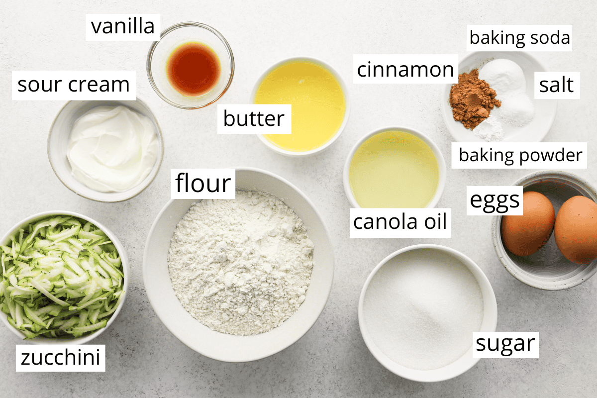 the ingredients for this Zucchini Cake recipe in bowls and labeled like sugar, flour, zucchini, eggs, butter, etc. 