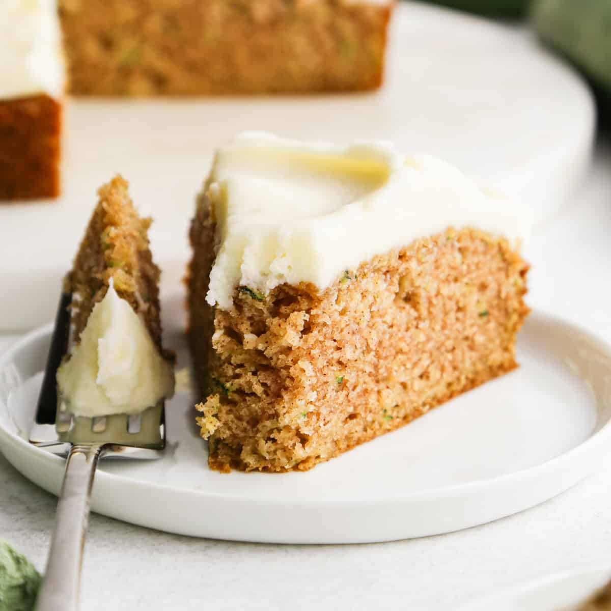 a slice of Zucchini Cake with cream cheese frosting on a white plate, a fork has taken a bite out of the front of the cake and is resting next to the cake on the plate. The rest of the zucchini cake is in the background