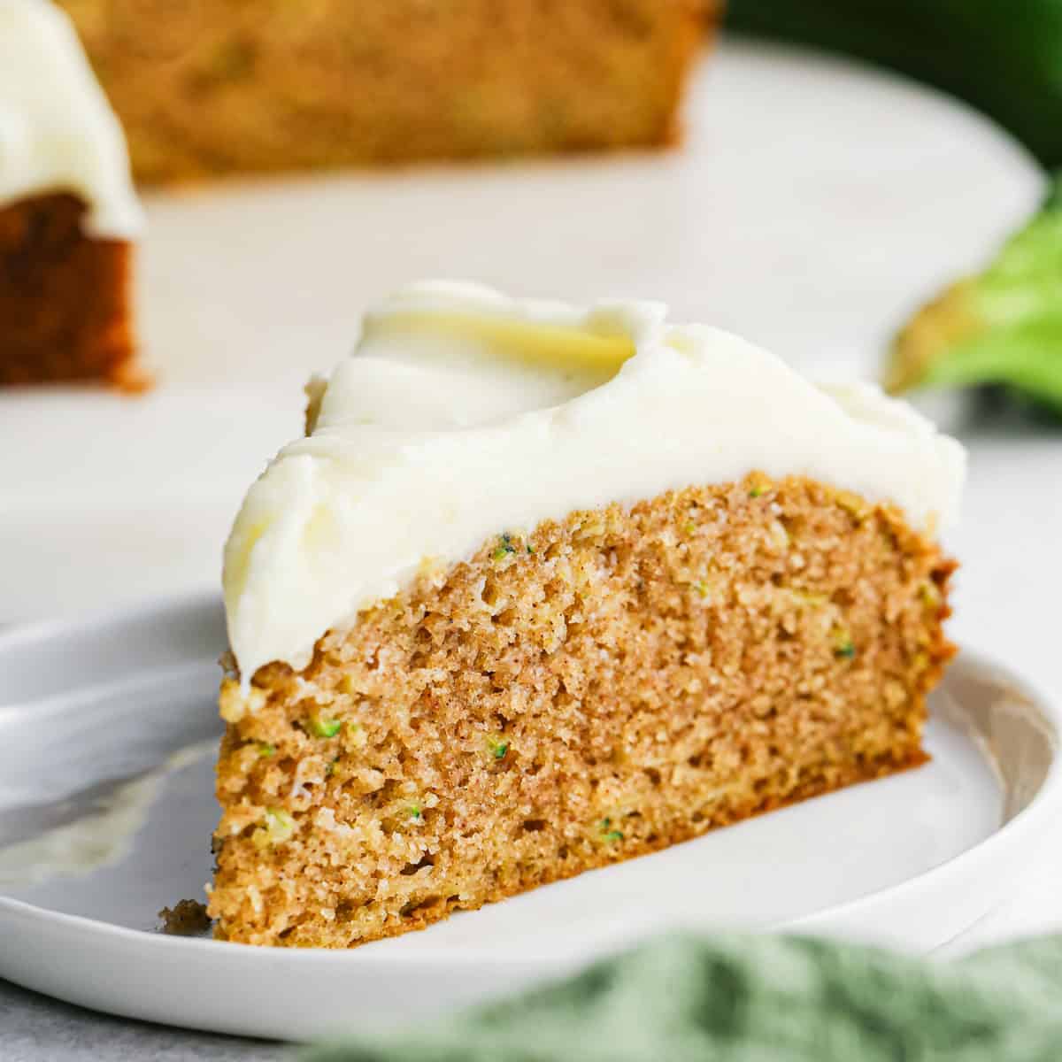 a slice of Zucchini Cake with cream cheese frosting on a white plate with the rest of the cake and a zucchini in the background