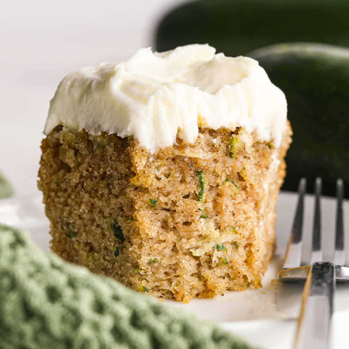 a piece of Zucchini Cake with cream cheese frosting on top. There is a bite taken out of it, a fork resting on the plate and zucchinis in the background