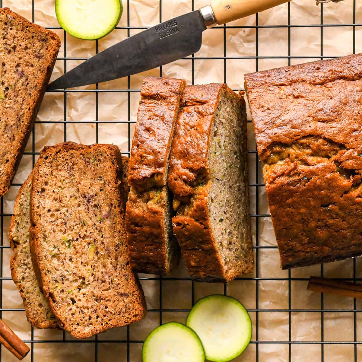 a loaf of Zucchini Banana Bread on a wire cooling rack on parchment paper with 5 slices cut out of it with sliced zucchini and a cinnamon stick nearby