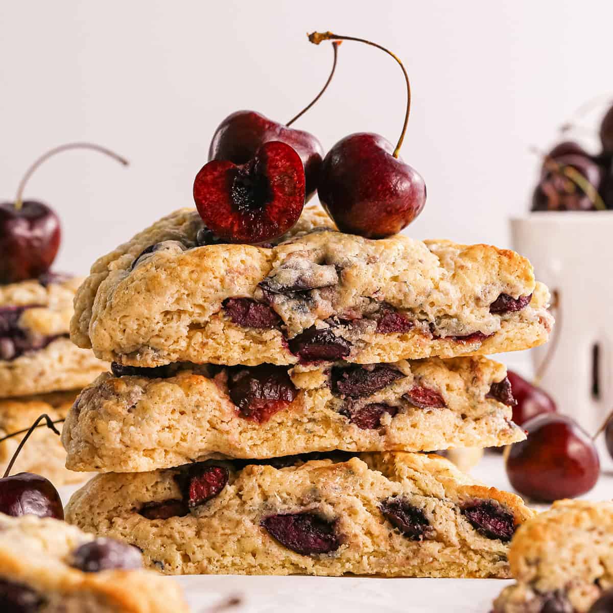 une pile de 3 scones aux cerises avec des cerises sur le dessus, entourée d'autres scones aux cerises