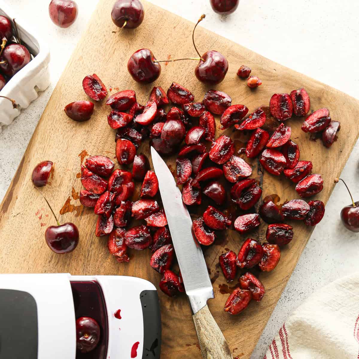 chopped cherries on a wood cutting board with a knife and the cherry pitter in the bottom lefthand corner 