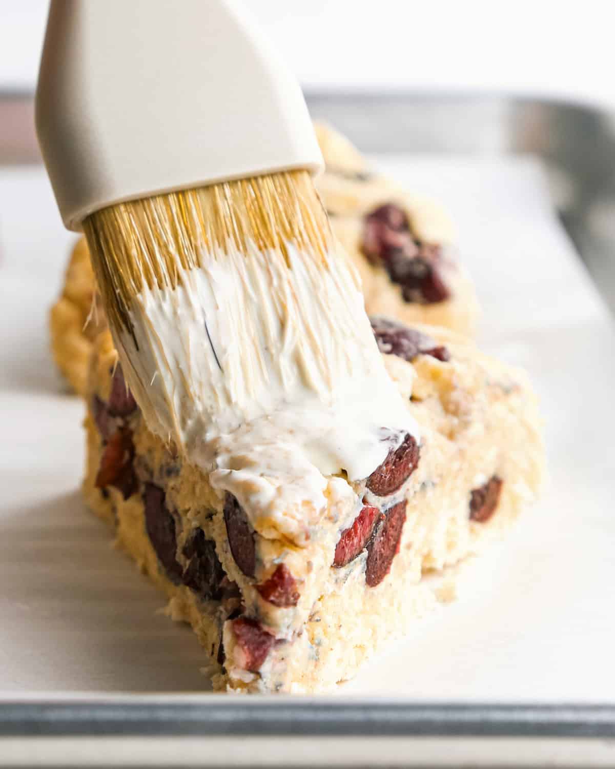 heavy cream being brushed on top of an unbaked cherry scone on a baking sheet lined with parchment paper