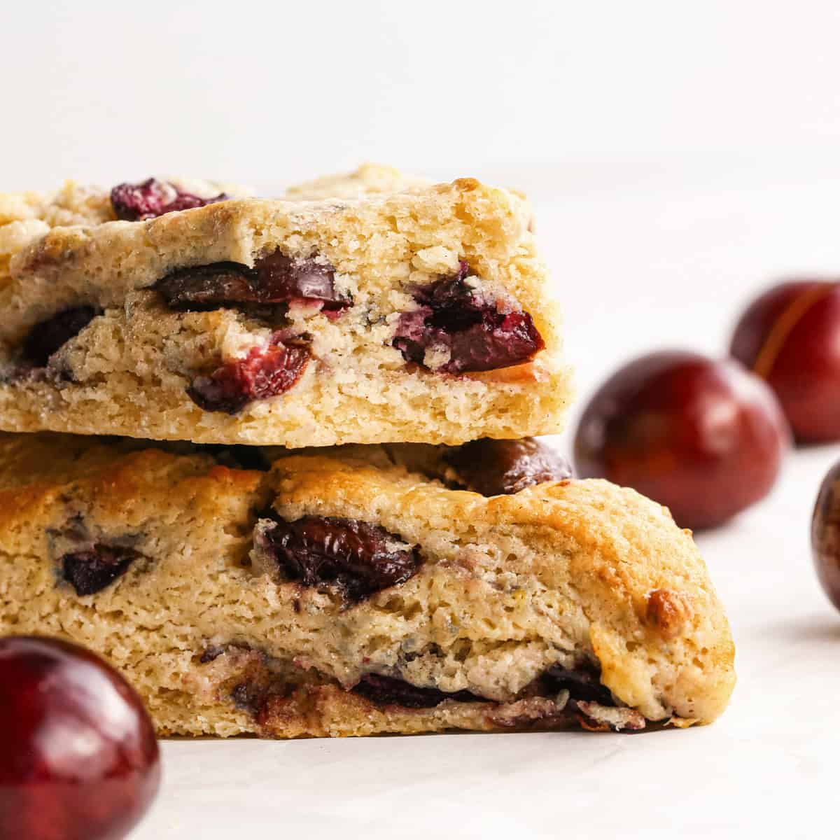 two cherry scones stacked on each other with cherries in the background, the top scone has a bite taken out of it