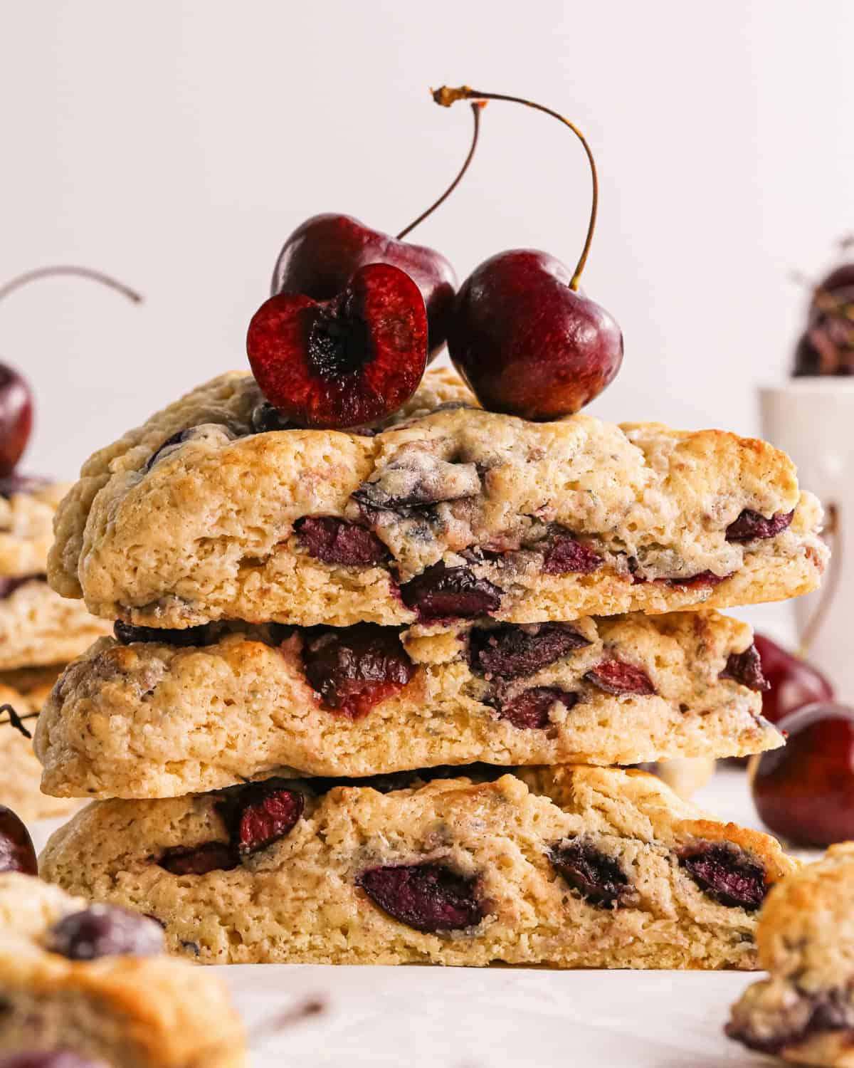 a stack of 3 cherry scones with 3 cherries on top, surrounded by other cherry scones and a bowl of cherries in the background