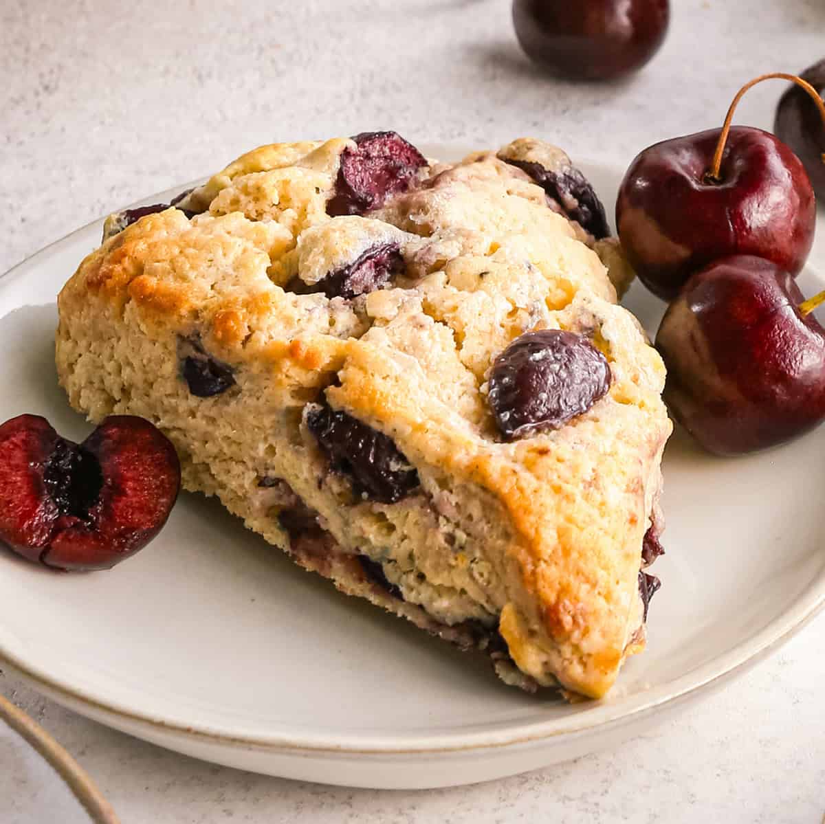 a cherry scone on a plate with two whole cherries and one half of a cherry on a gray background