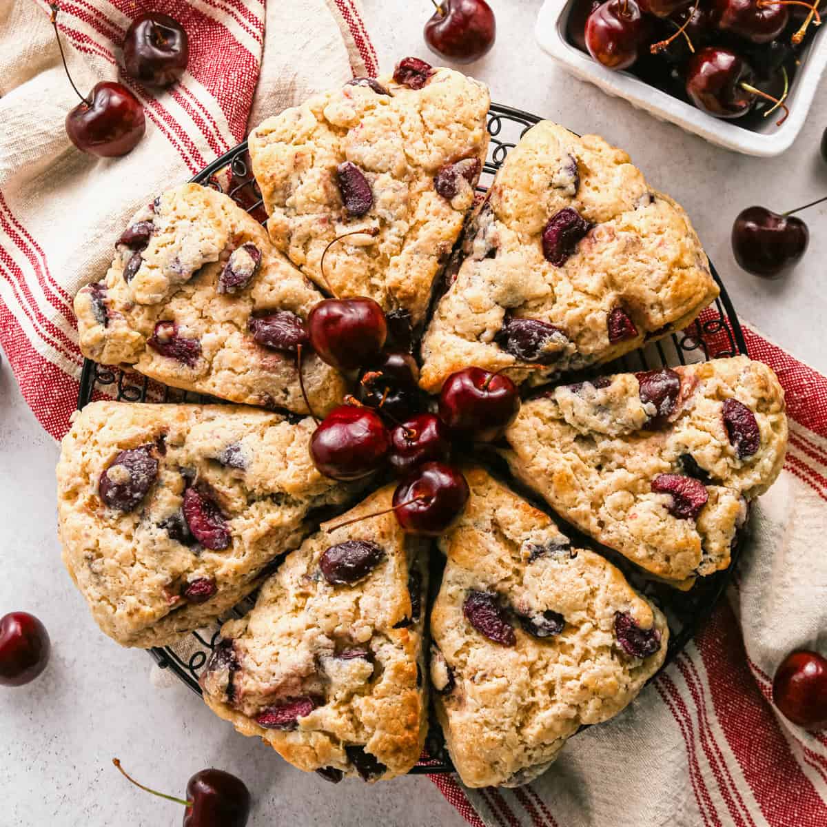 7 cherry scones arranged in a circle on a circular wire cooling rack with a red and cream towel and cherries surrounding them
