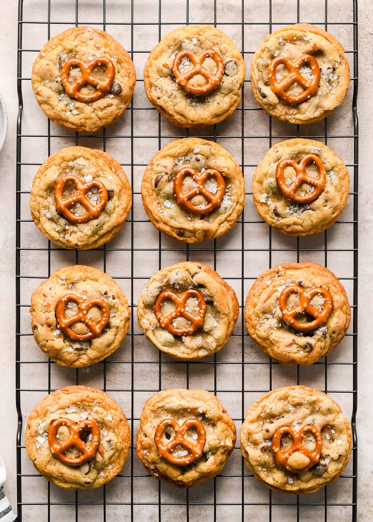 12 Chocolate Chip Pretzel Cookies on a wire cooling rack each topped with a mini pretzel and flaky sea salt