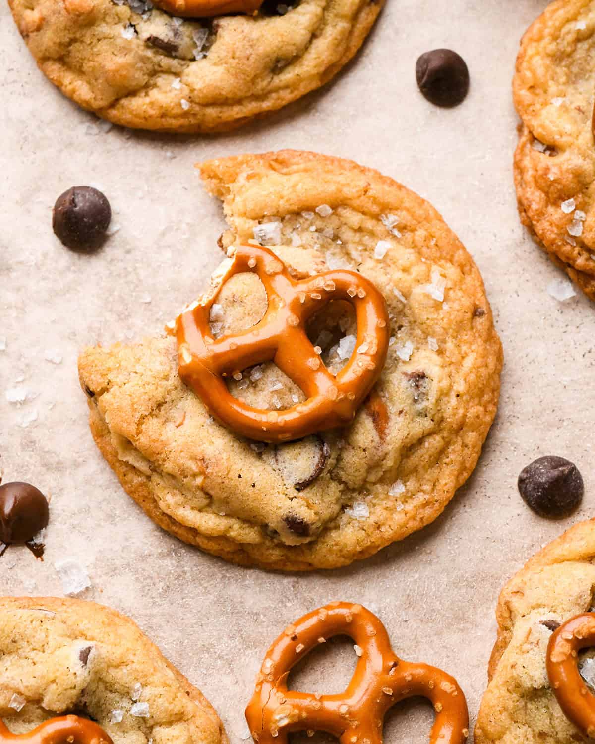 a chocolate chip pretzel cookie with a bite taken out of it topped with a mini pretzel and a sprinkle of flaky sea salt. surrounded by chocolate chips and pretzels and more cookies