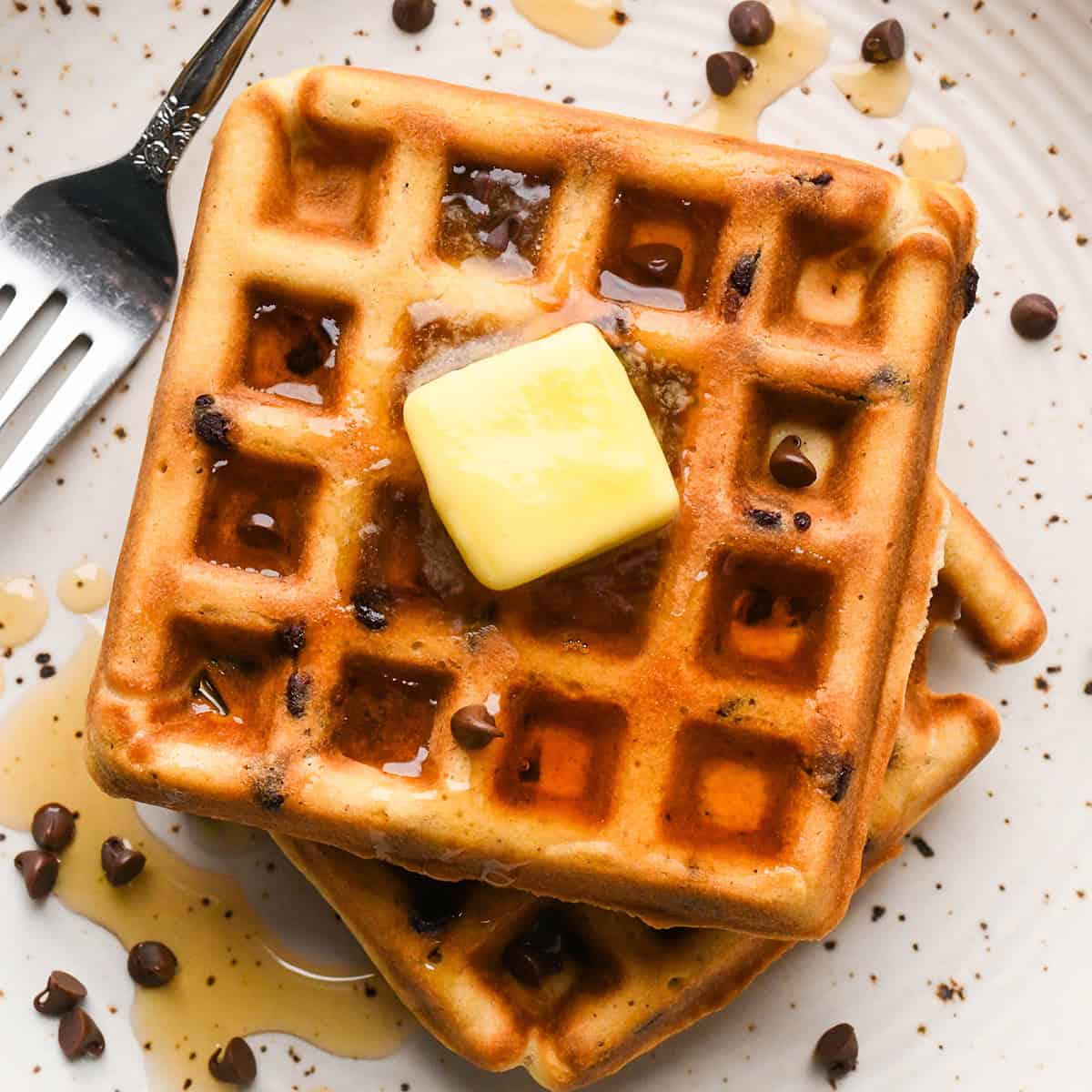 a stack of 2 Chocolate Chip Waffles on a white plate topped with butter and syrup, with mini chocolate chips and a fork on the plate