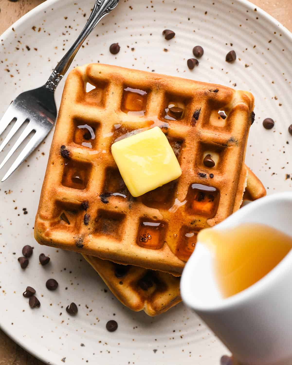 syrup being poured over a stack of two Chocolate Chip Waffles with butter on top on a white plate with mini chocolate chips and a fork