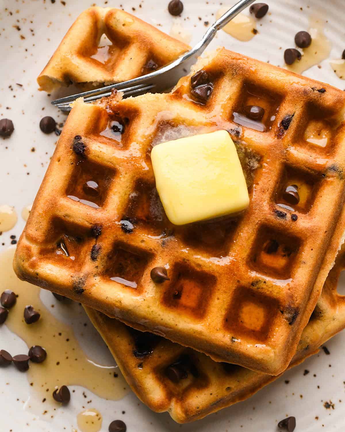 two Chocolate Chip Waffles on a plate with a fork taking a bite, topped with butter and syrup and chocolate chips