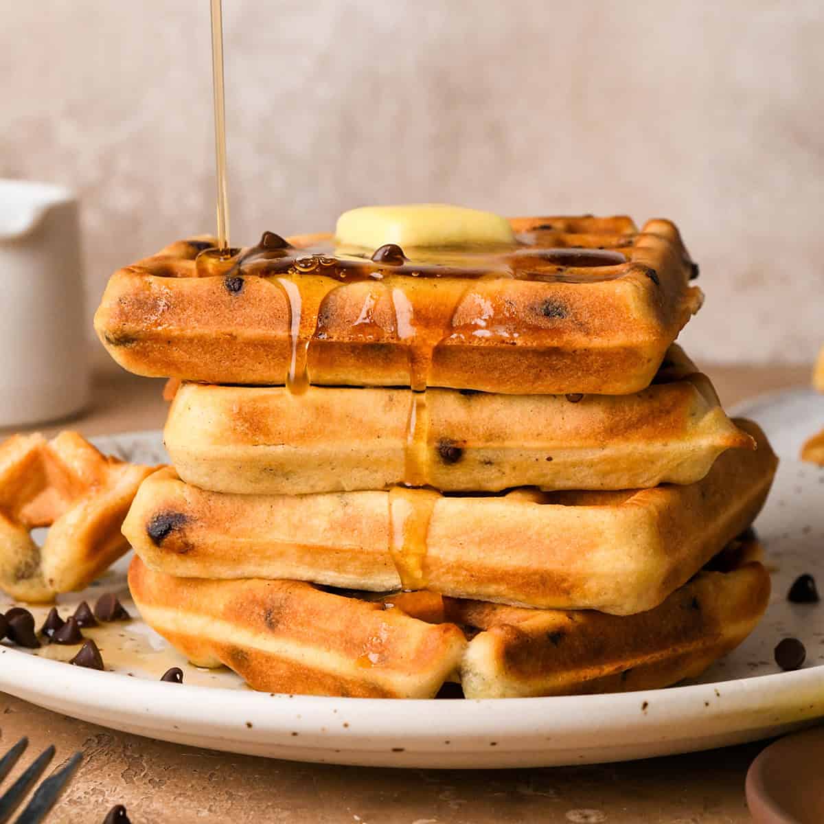 syrup being poured over a stack of 4 Chocolate Chip Waffles on a white plate with mini chocolate chips on the plate