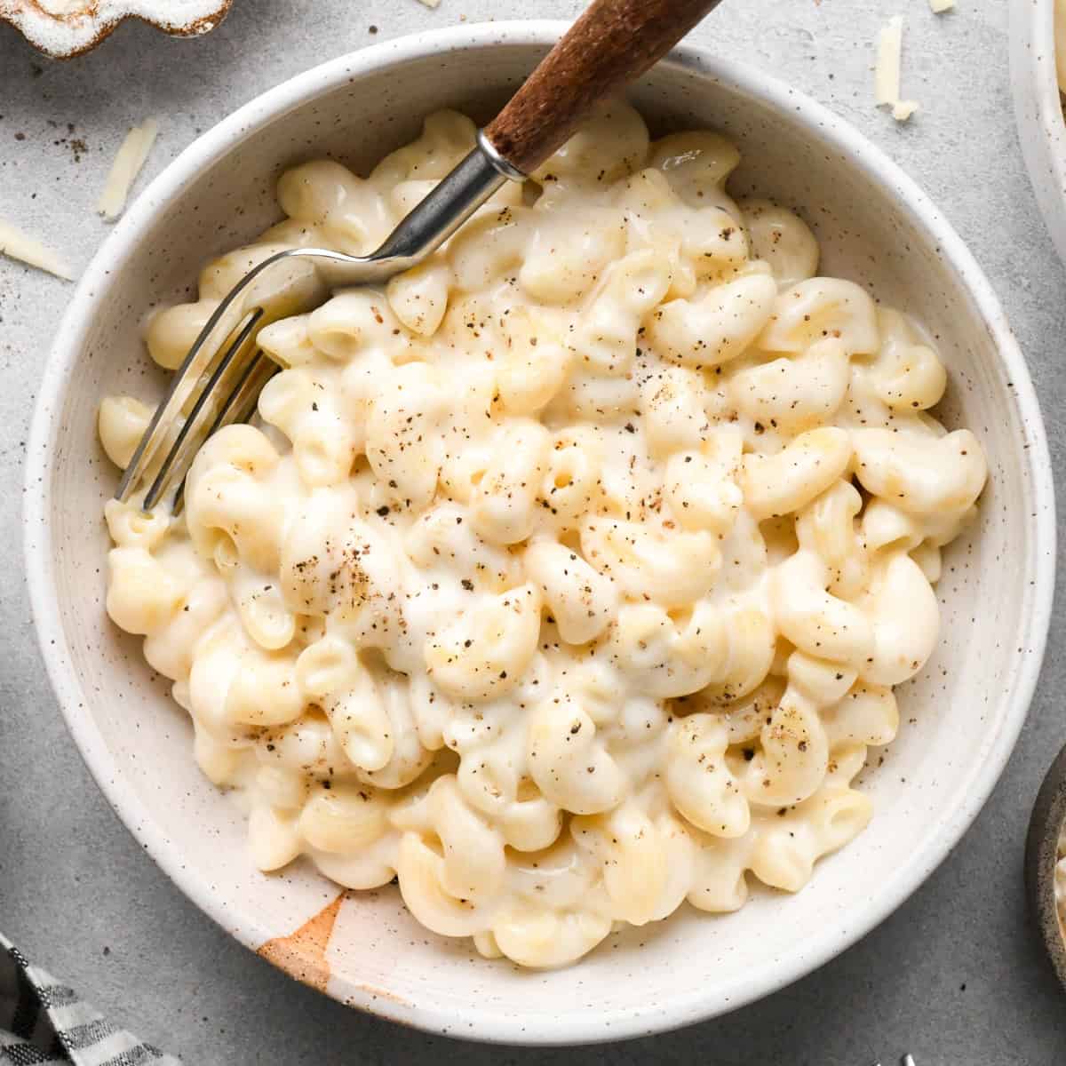 cottage cheese mac and cheese in a ceramic bowl with a fork garnished with freshly ground black pepper with shredded cheese on the gray surface it's on