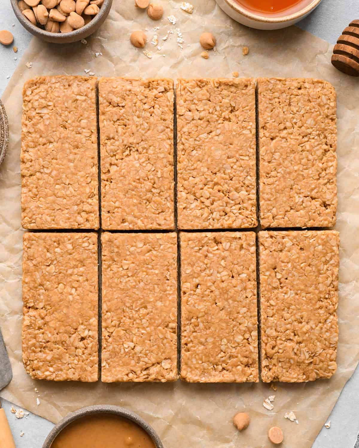 Peanut Butter Granola Bars cut into 8 rectangles on parchment paper surrounded by peanut butter chips and oats