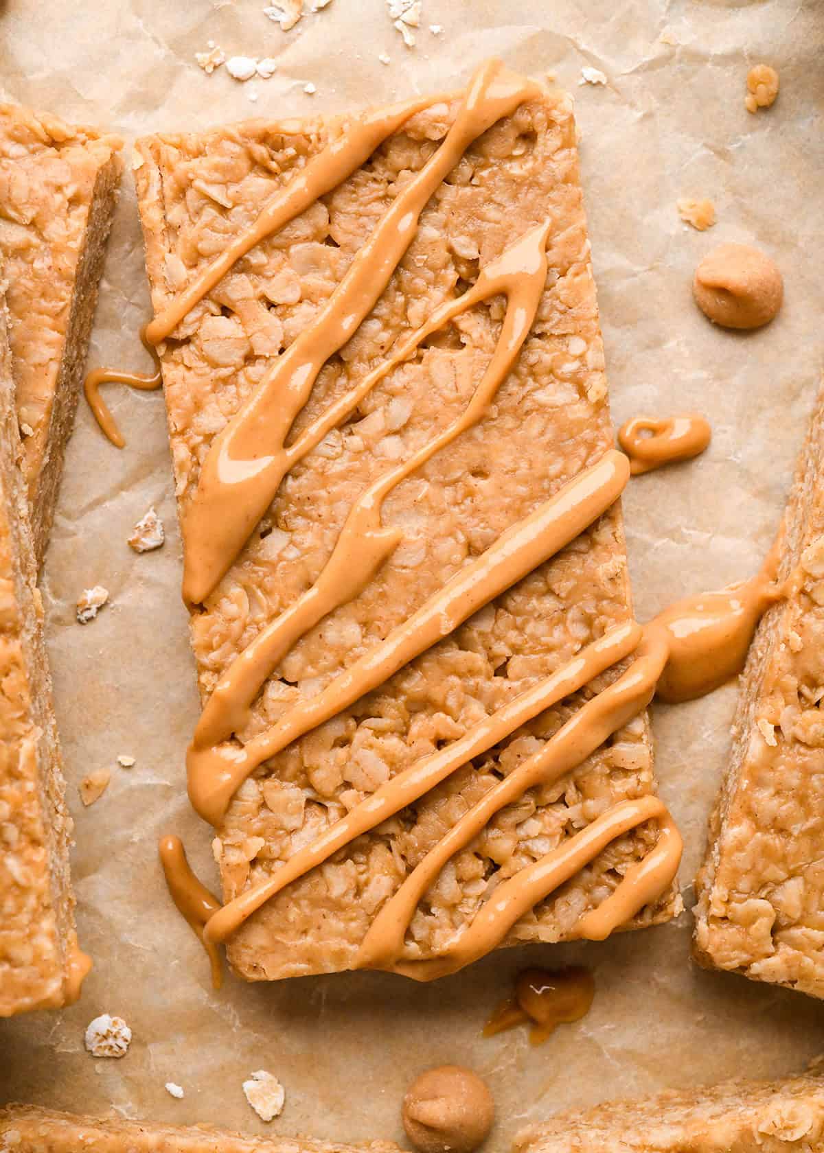 peanut butter granola bar with a peanut butter drizzle on top on parchment paper surrounded by peanut butter chips and other peanut butter granola bars 