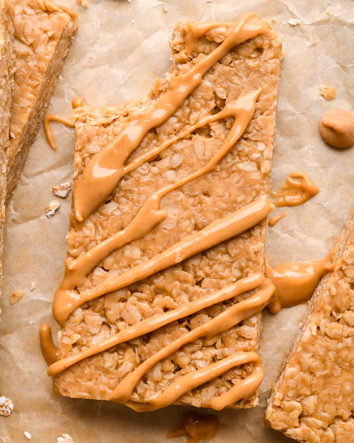a Peanut Butter Granola Bar with a peanut butter drizzle and bite taken out of it on parchment paper surrounded by more granola bars and peanut butter chips